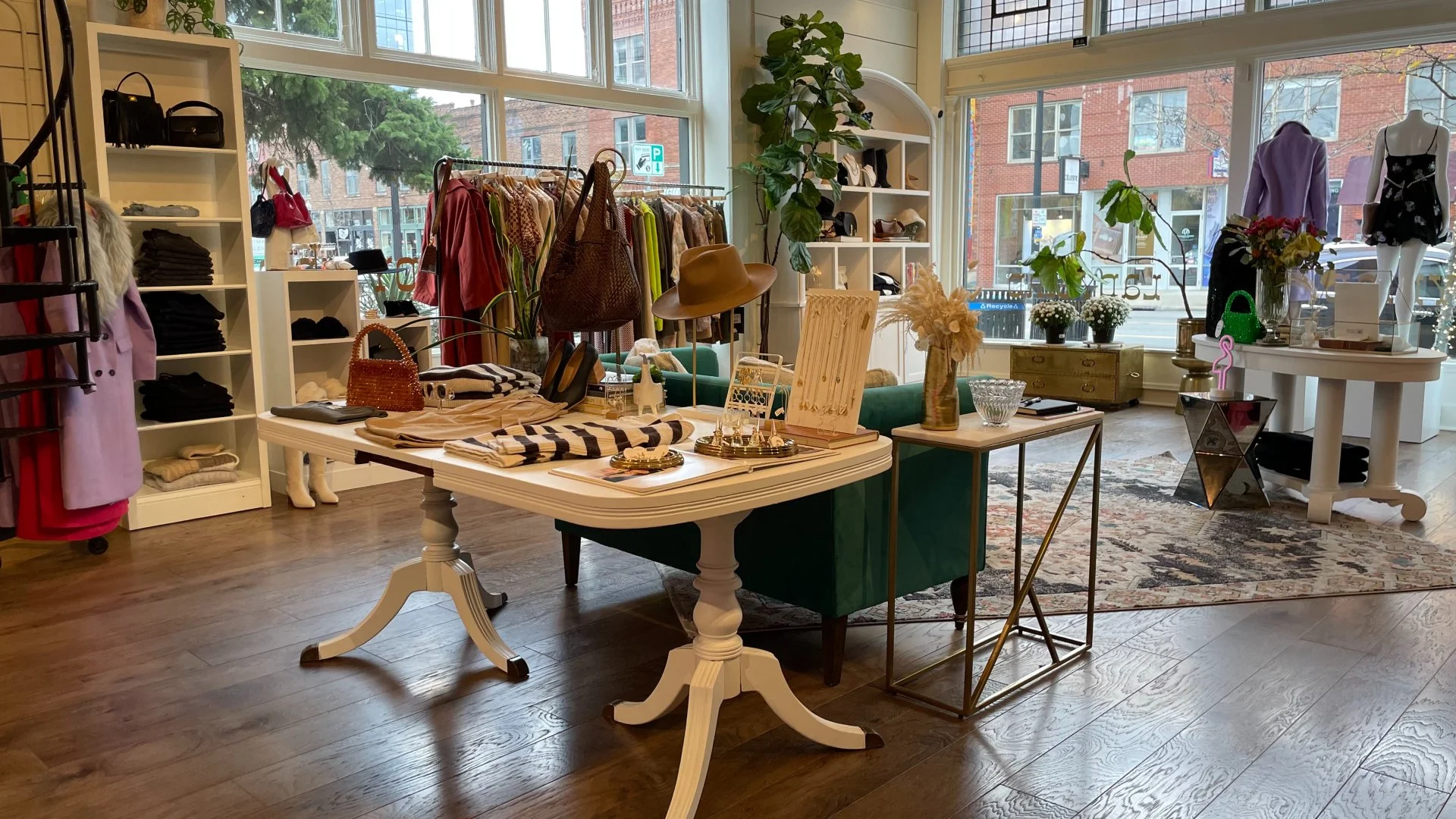 Interior of a boutique clothing store with shelves of folded clothes, racks of hanging garments, and display tables featuring accessories, hats, and jewelry. Large windows let in natural light and reveal city buildings outside.