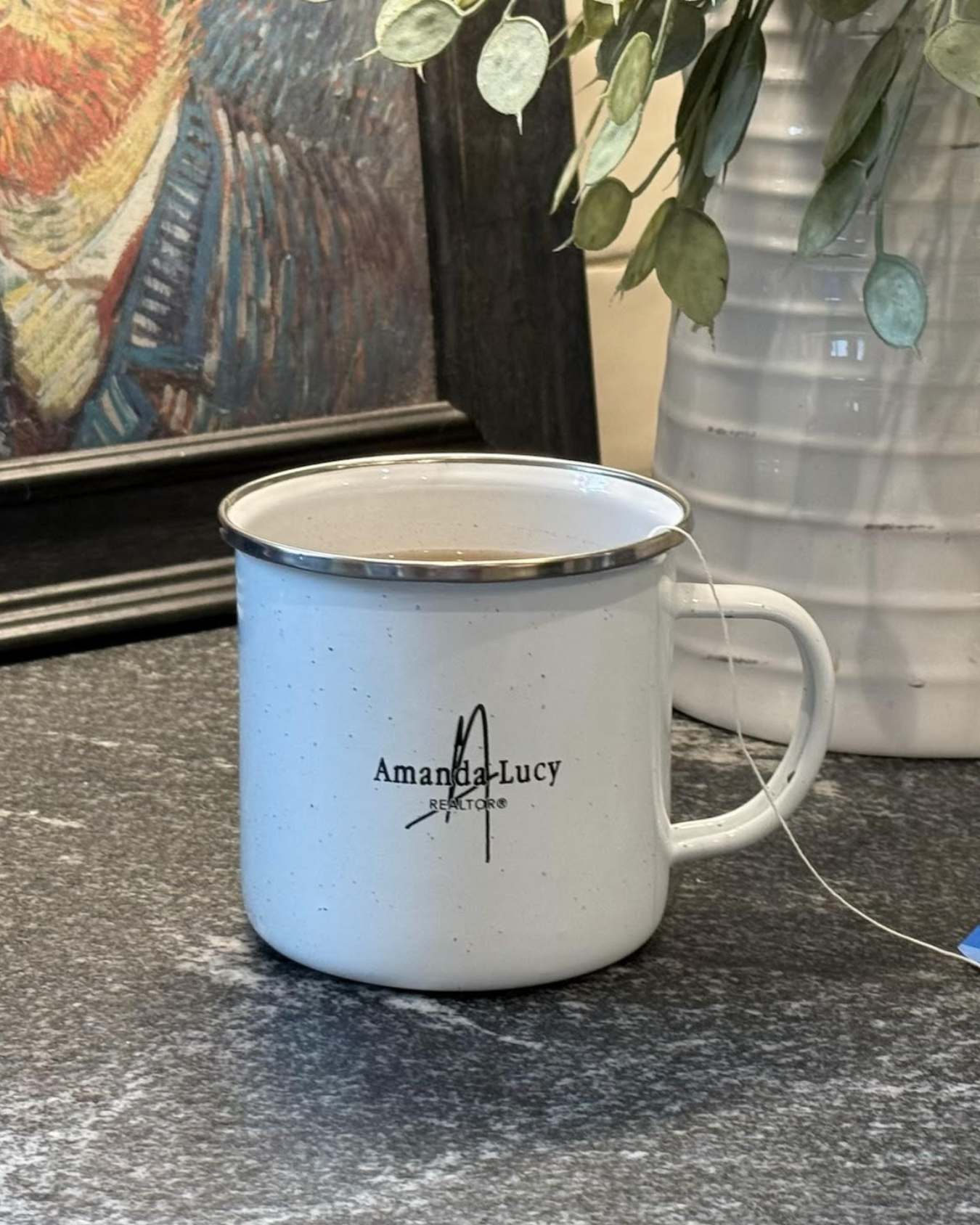 White enamel mug with "Amanda Lucy" text sits on a countertop next to a vase and a framed artwork in the background for people after buying a home with a local Realtor