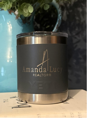 A stainless steel tumbler with "Amanda Lucy Realtor" printed on it sits on a closed book next to a blue vase.
