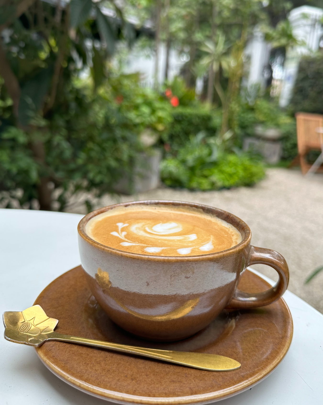 A cup of latte with latte art sits on a brown saucer with a gold spoon, outdoors in a garden setting to show selling a home with a Realtor in Columbus