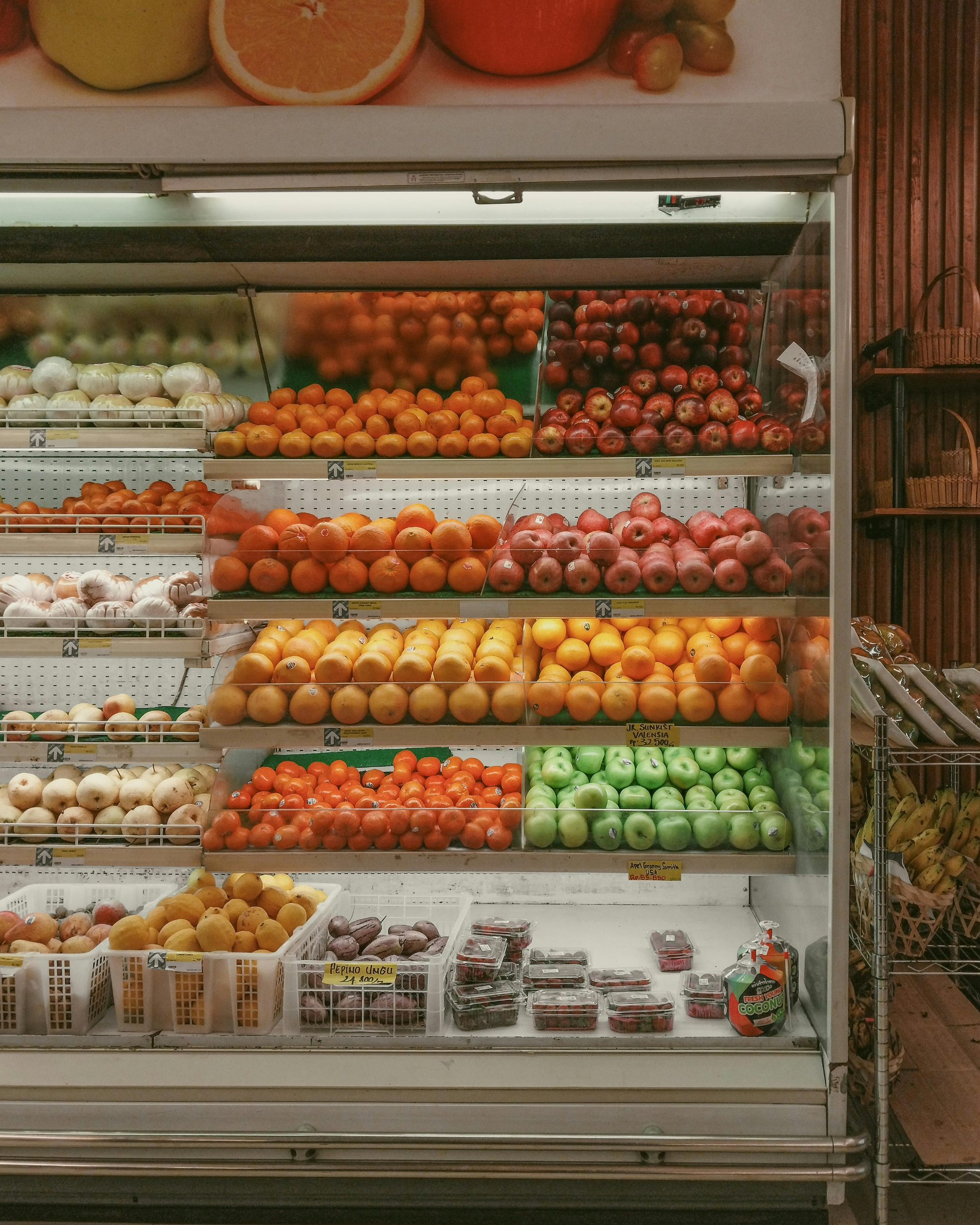 Supermarket refrigerator shelves stocked with apples, oranges, tomatoes, berries, and other assorted fruits.