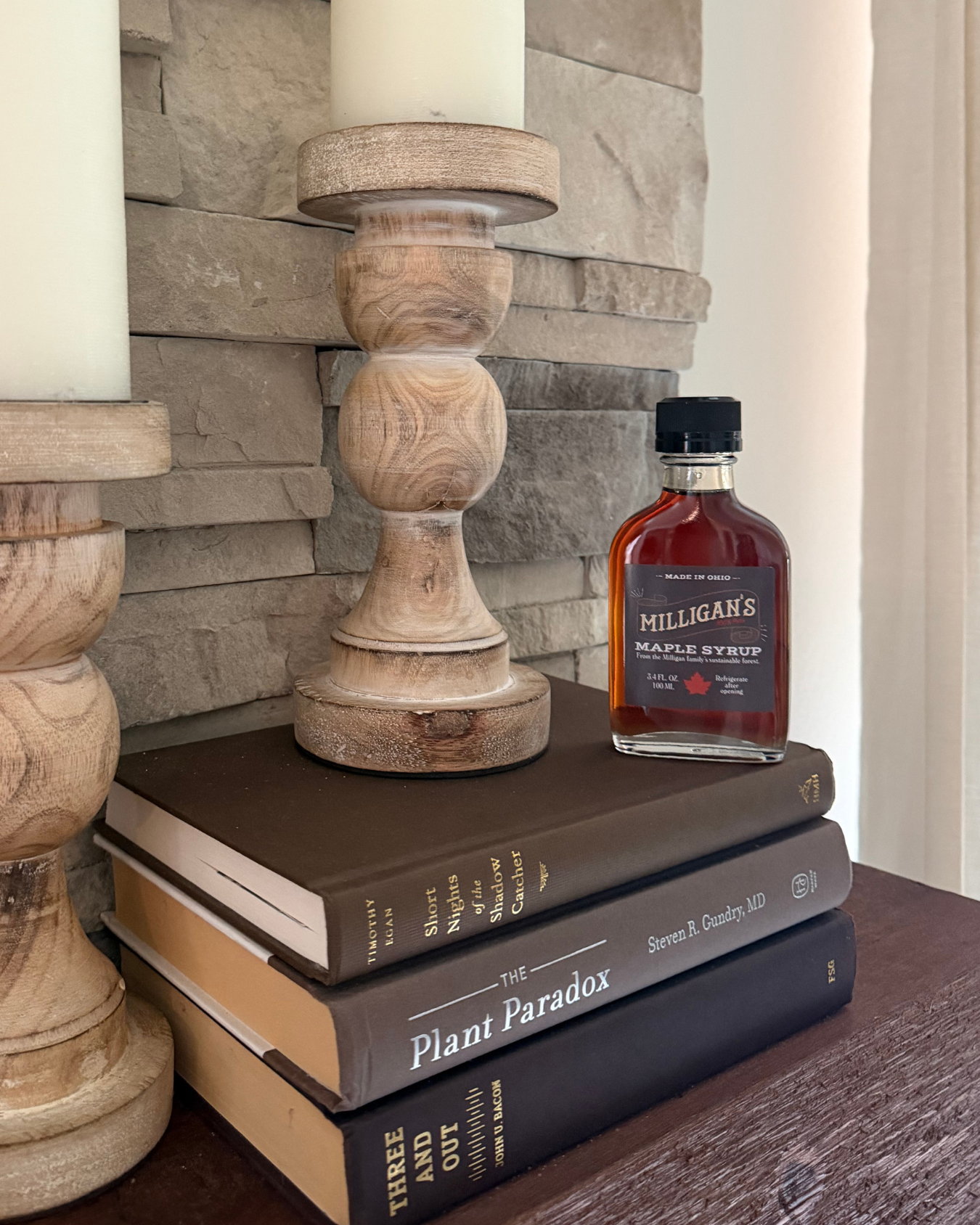 A bottle of maple syrup on stacked books beside two wooden candlesticks in front of a stone wall for Cutler Real Estate in Columbus Ohio