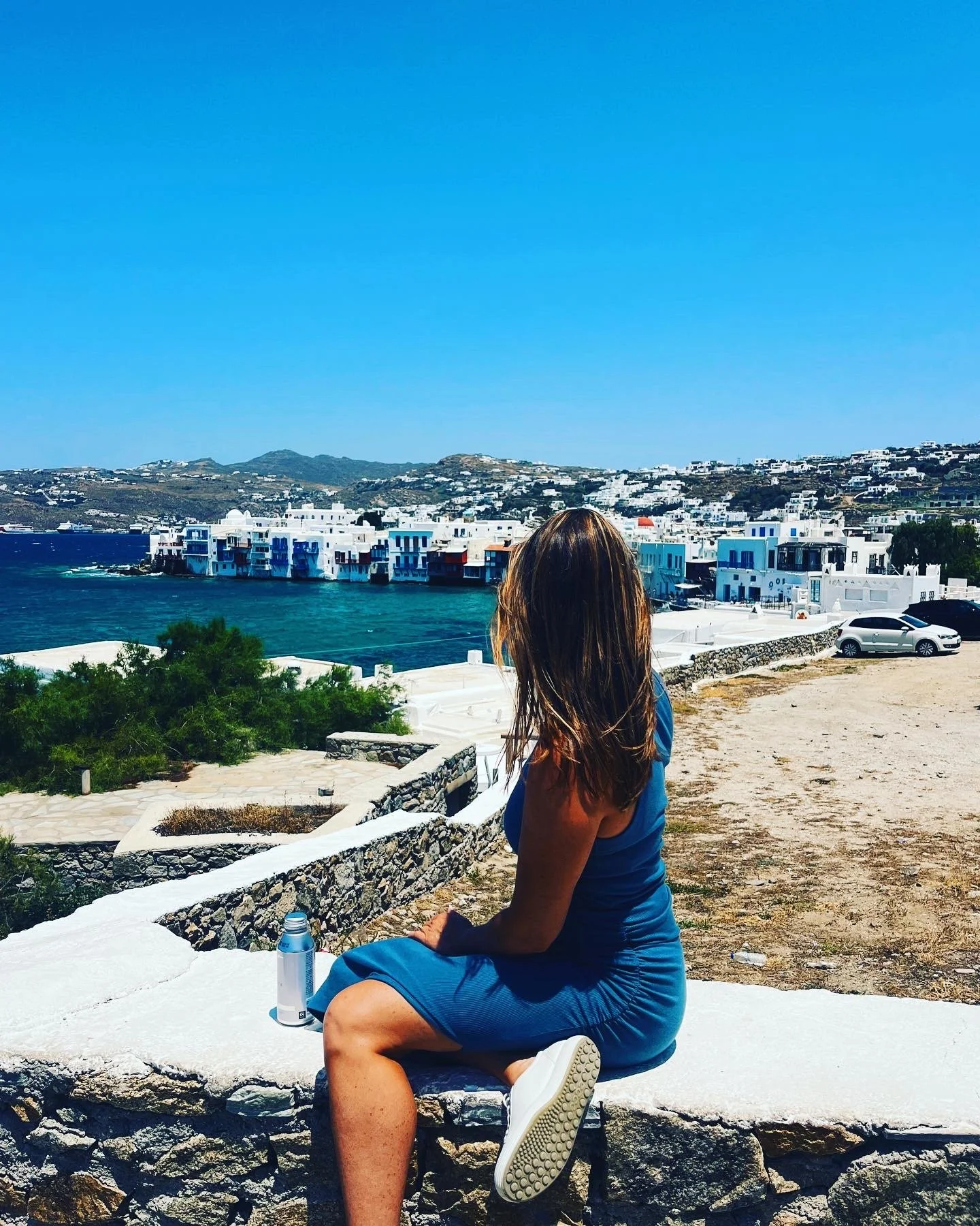 Columbus Ohio relocation Realtor, Amanda Lucy in a blue dress sits on a white stone wall overlooking coastal buildings and blue sea under a clear sky.