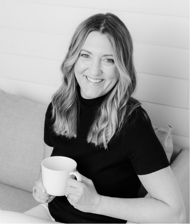 Amanda Lucy, A Columbus Ohio Realtor,  with long hair in a black top sits on a couch, smiling and holding a mug.