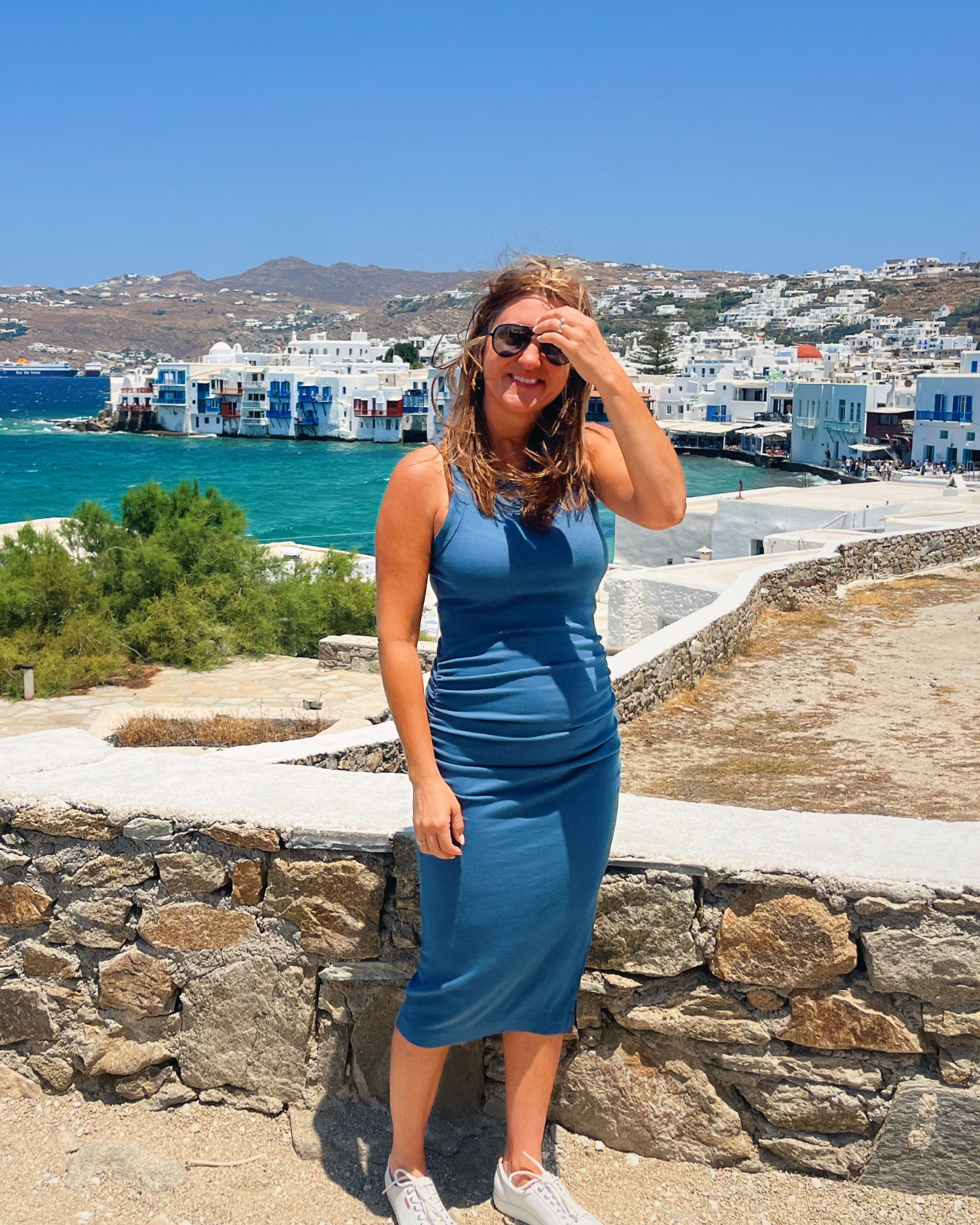 Top Realtor in Columbus Ohio, Amanda Lucy in a blue dress standing by a stone wall with coastal buildings and blue sea in the background on a sunny day.