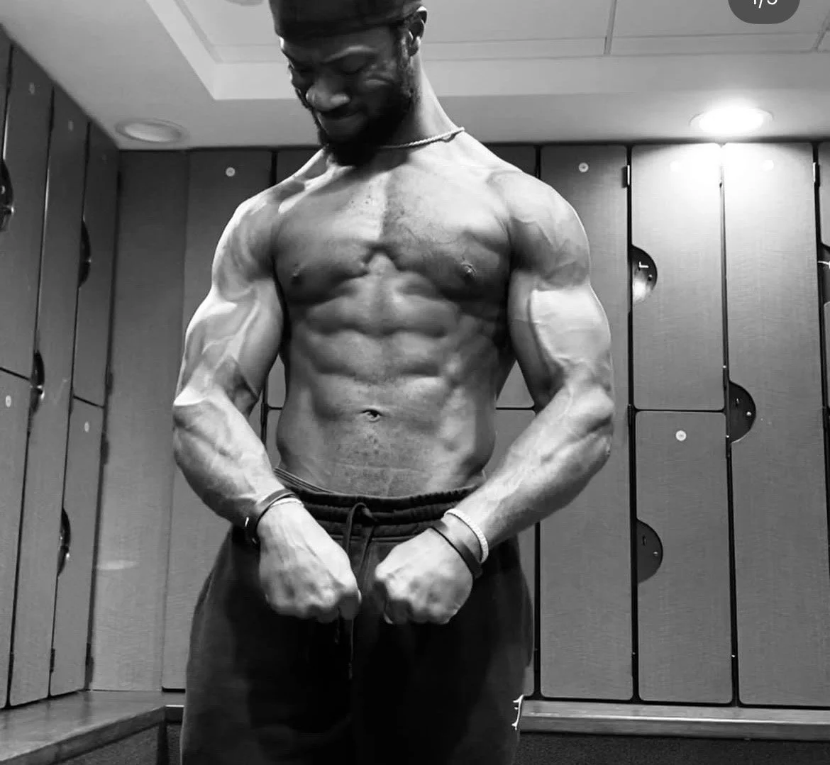 A muscular man flexing his biceps in a gym locker room, showing defined abs and arm muscles.
