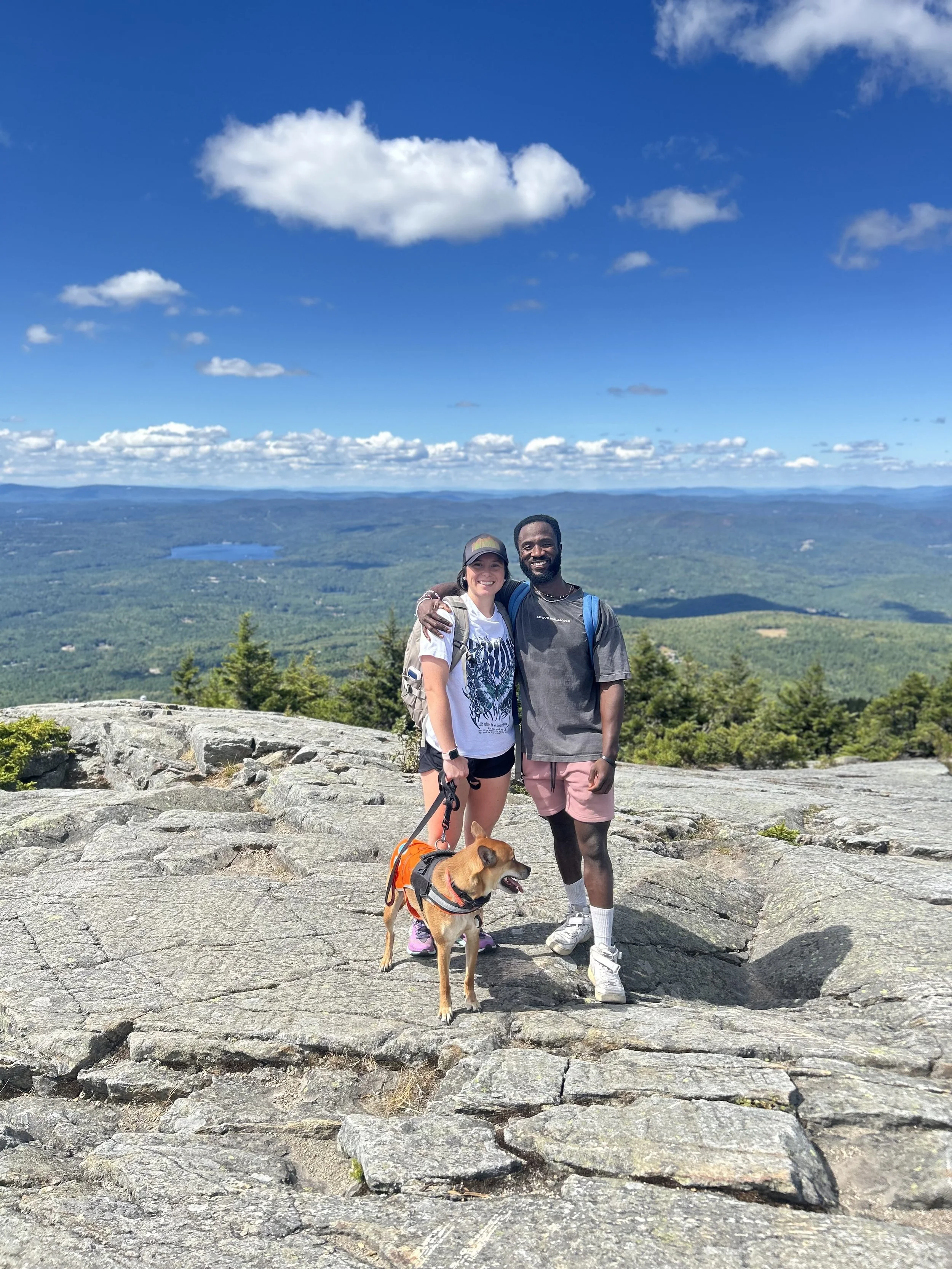 A smiling couple with their dog on a rocky mountaintop with a scenic view of trees, lakes, and blue sky with clouds in the background.