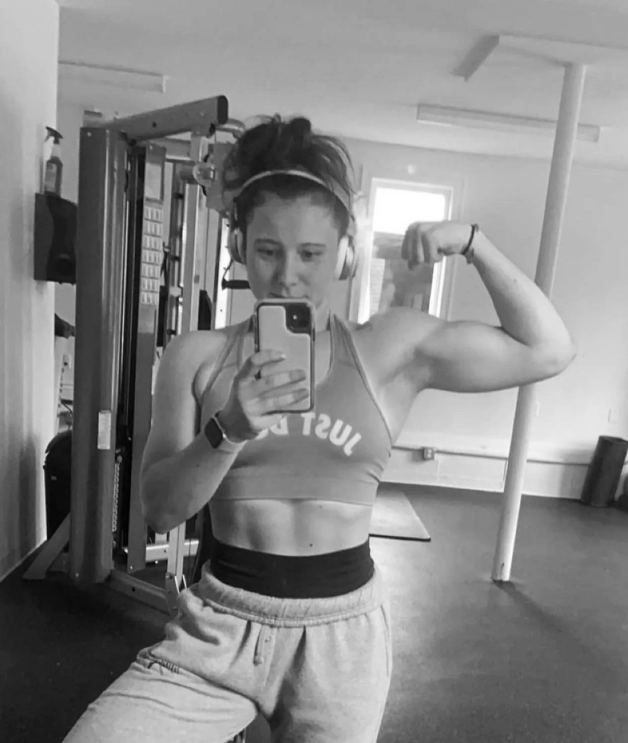 A woman at the gym taking a mirror selfie, flexing her right bicep, wearing headphones, a sports bra, and sweatpants.