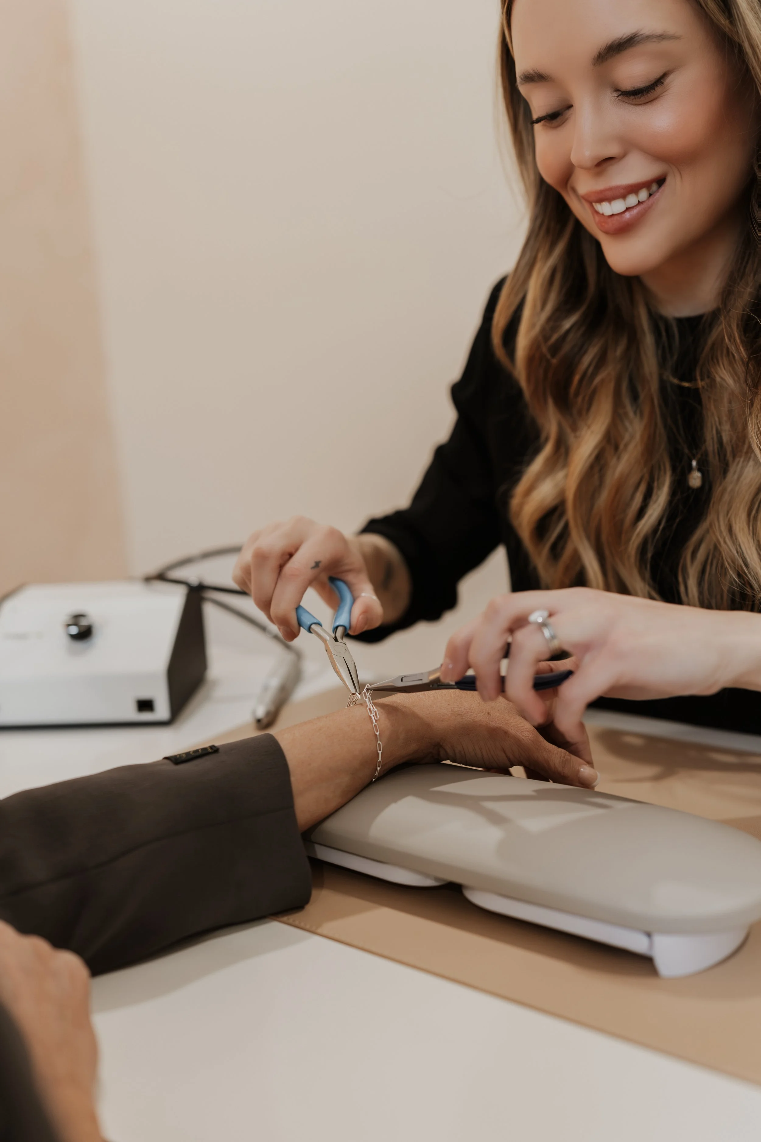 A woman with long wavy hair taking a patient's pulse with a pulse oximeter on an elderly person's wrist. The woman is smiling and using medical tools.