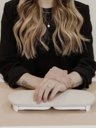 A woman with long, wavy hair wearing a black blazer and a necklace, resting her hands on a white gender-neutral scale on a beige surface.