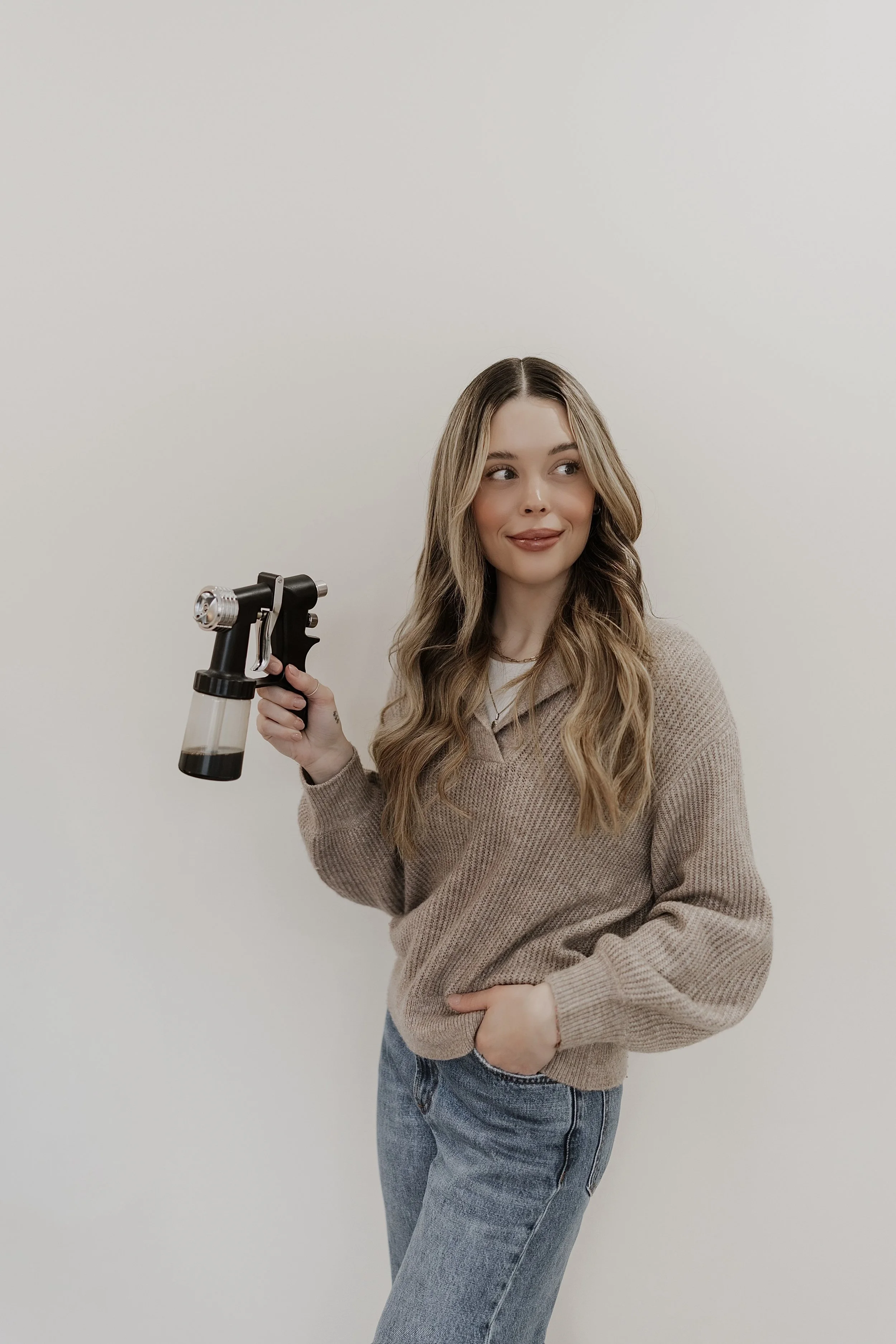 A young woman with long wavy hair and a beige sweater holds a small, black, and silver ray gun prop, standing against a plain white wall.
