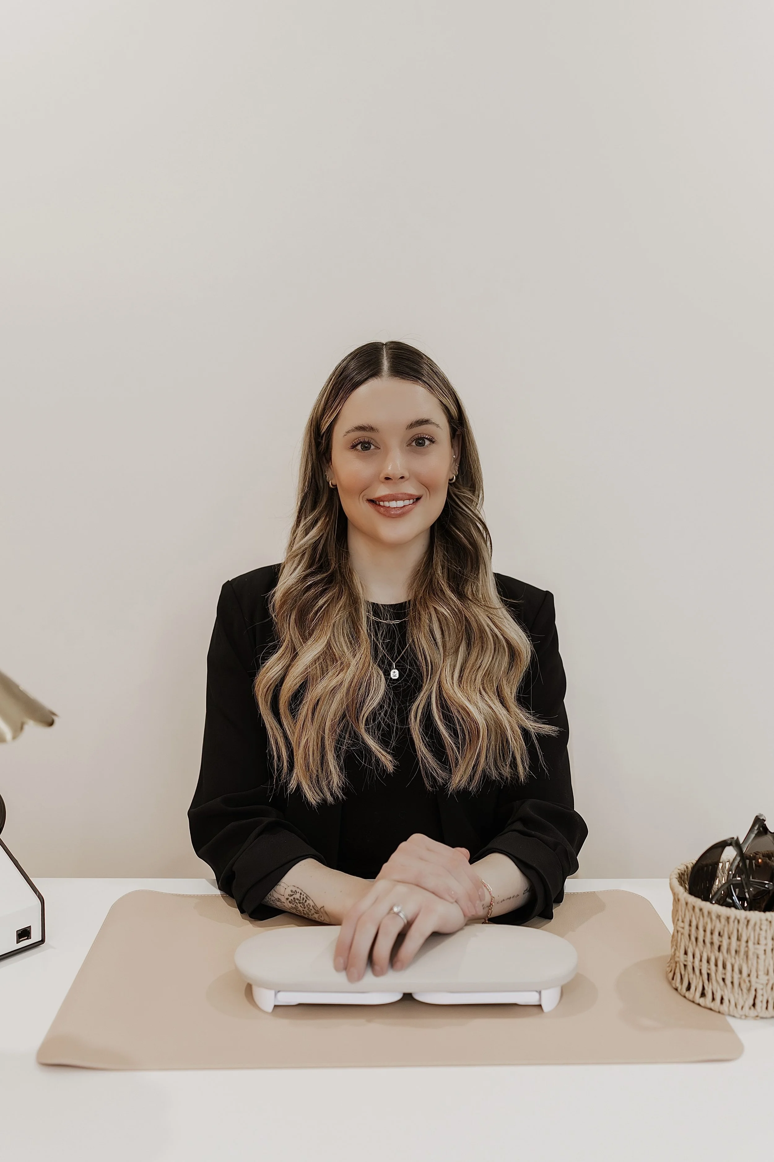 A woman with long wavy hair, wearing a black blazer, sitting at a white desk with her hands resting on a white stand, smiling, with a beige placemat and a small lamp on the left and a woven basket with black glasses on the right, against a plain white wall background.