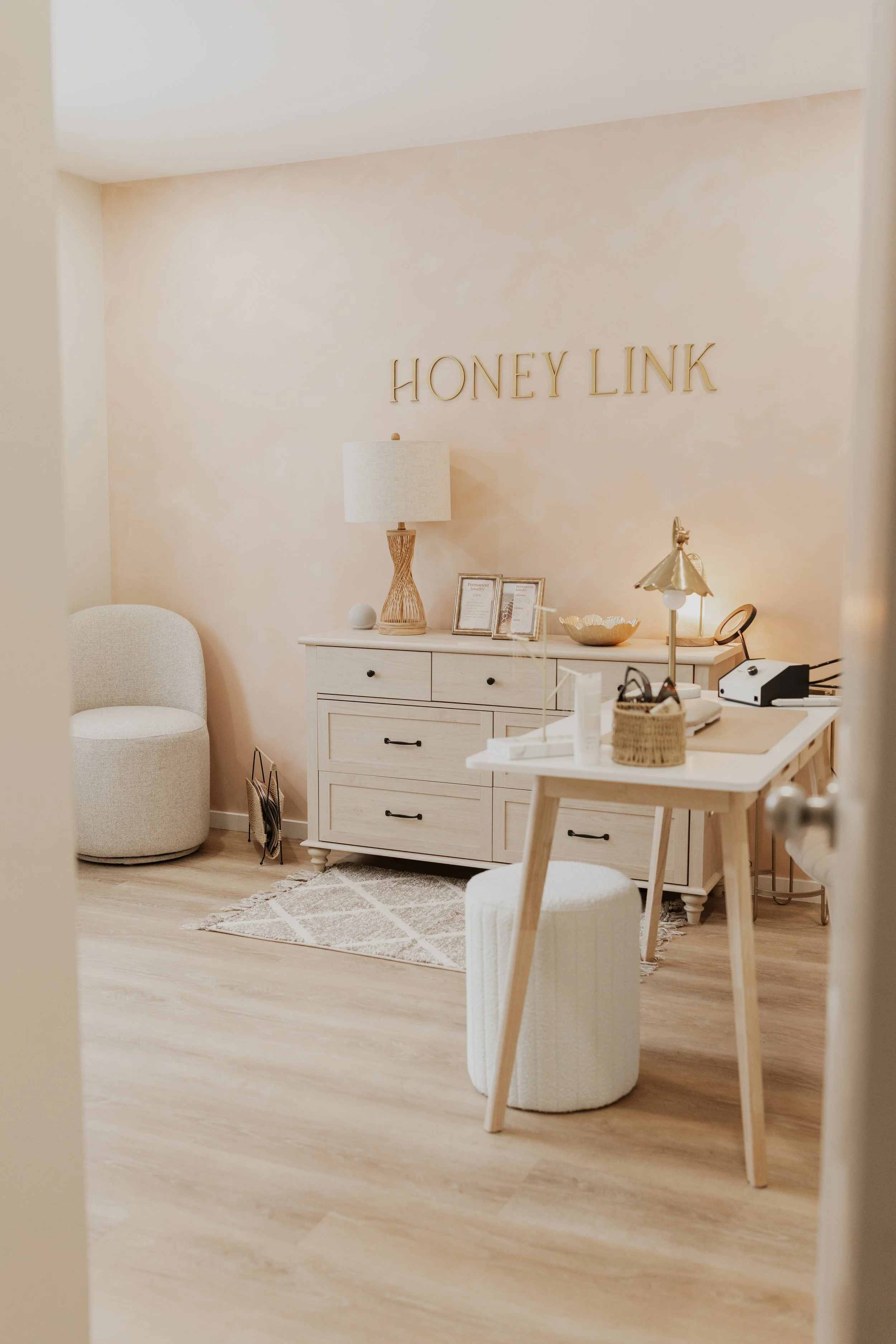 Interior of a cozy office with a white desk, a white upholstered stool, a beige armchair, a lamp, framed pictures, and decorative items, with the sign 'Honey Link' on the wall.