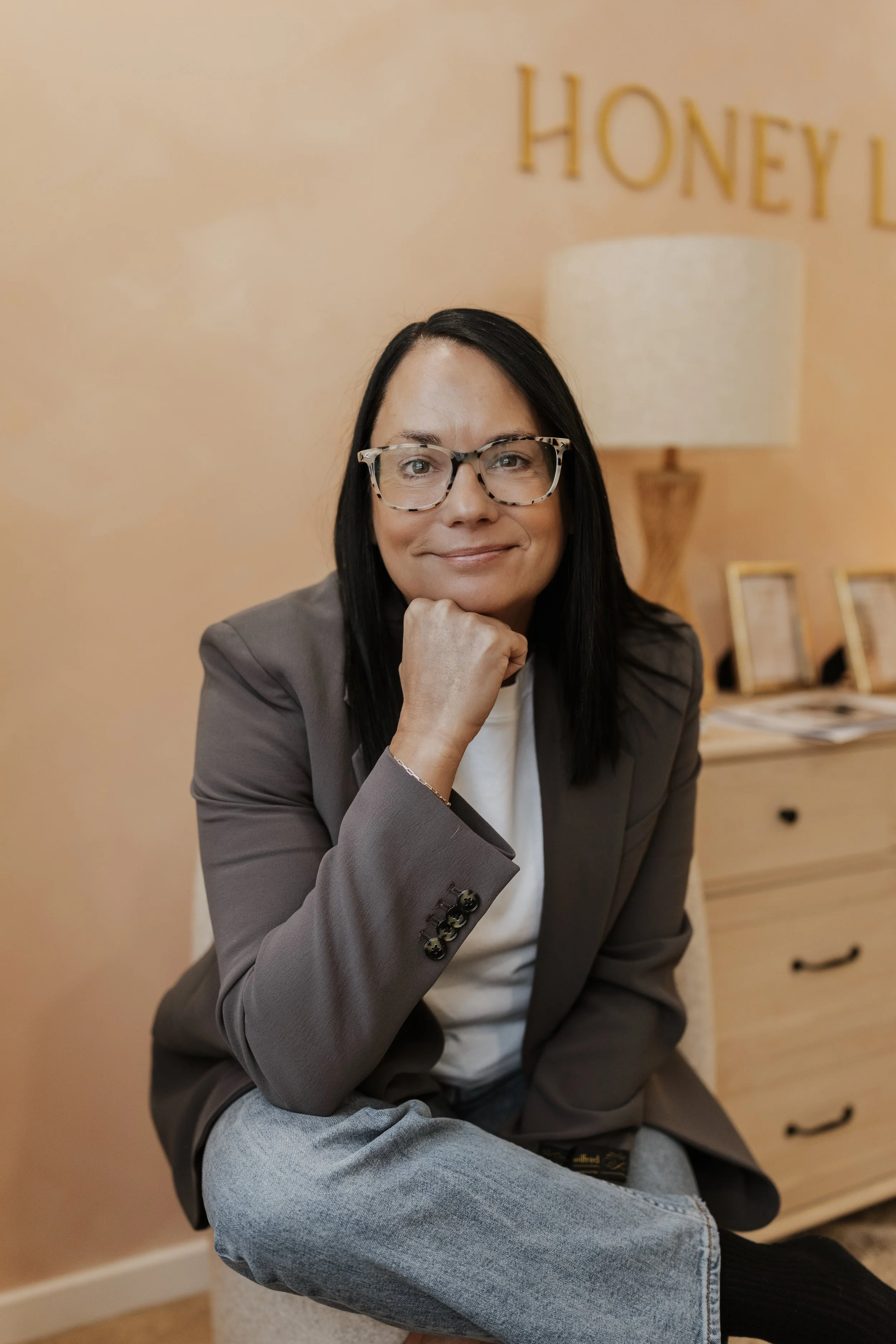 A woman with black hair and glasses, wearing a gray blazer and light-colored jeans, sitting indoors with her chin resting on her hand, smiling. Behind her is a beige wall with the word 'HONEY' partially visible, a wooden dresser, and a lamp.