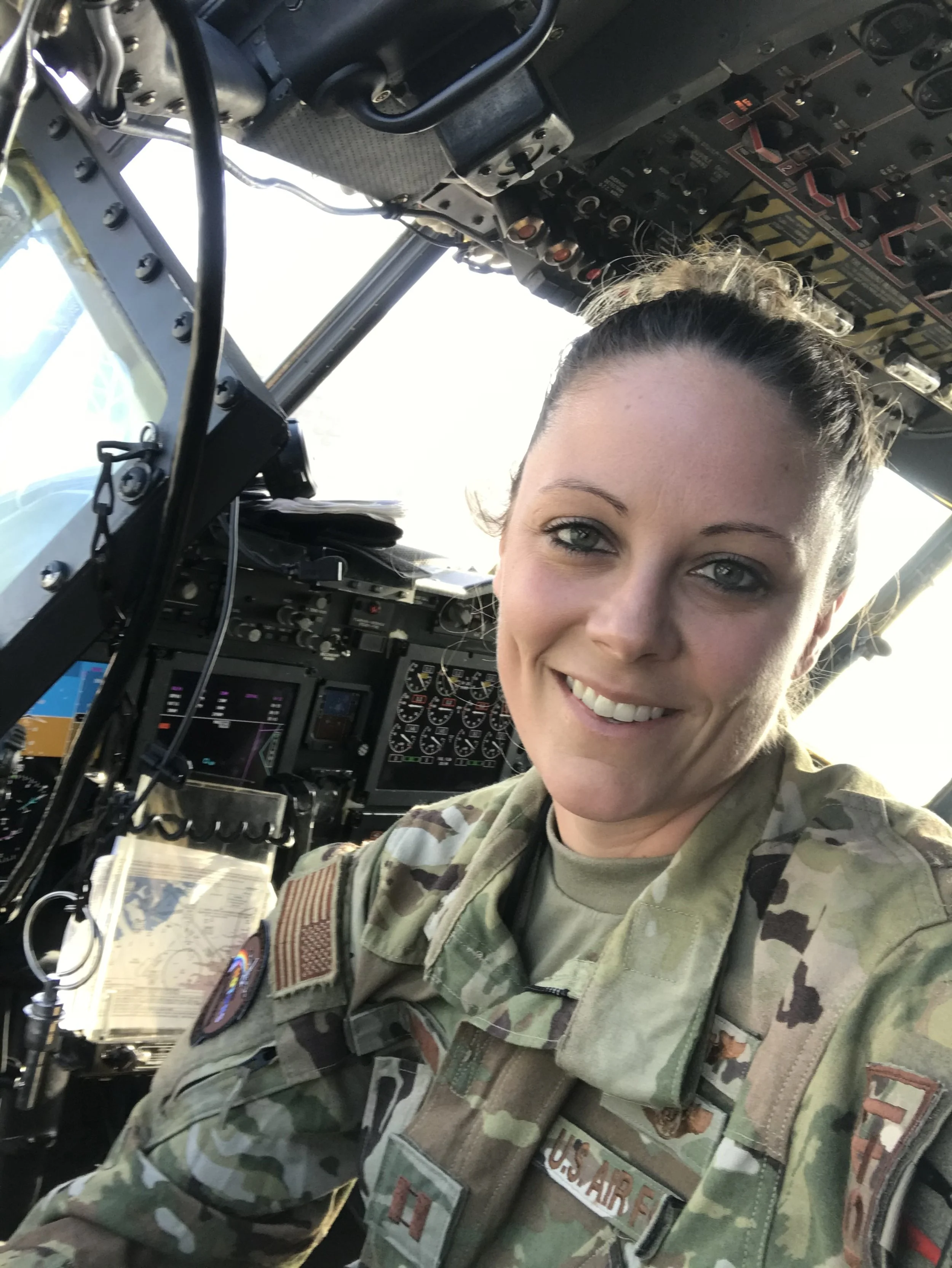 A woman in military uniform takes a selfie in the cockpit of an aircraft, smiling at the camera.