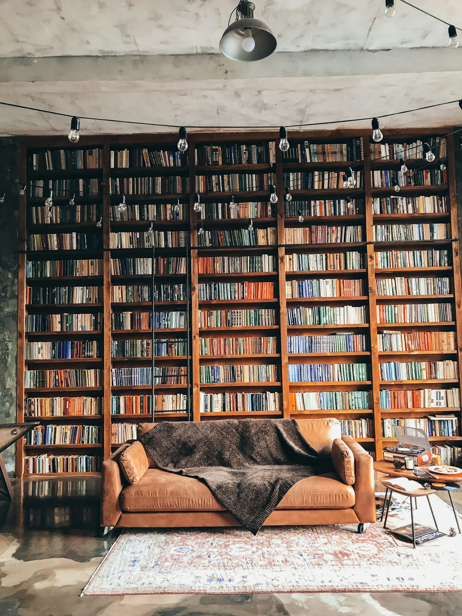 A cozy reading nook with a large wooden bookshelf filled with books, a tan velvet sofa with a dark throw blanket, a patterned rug, and a small table with books and decor items, in an industrial-style room with hanging string lights.