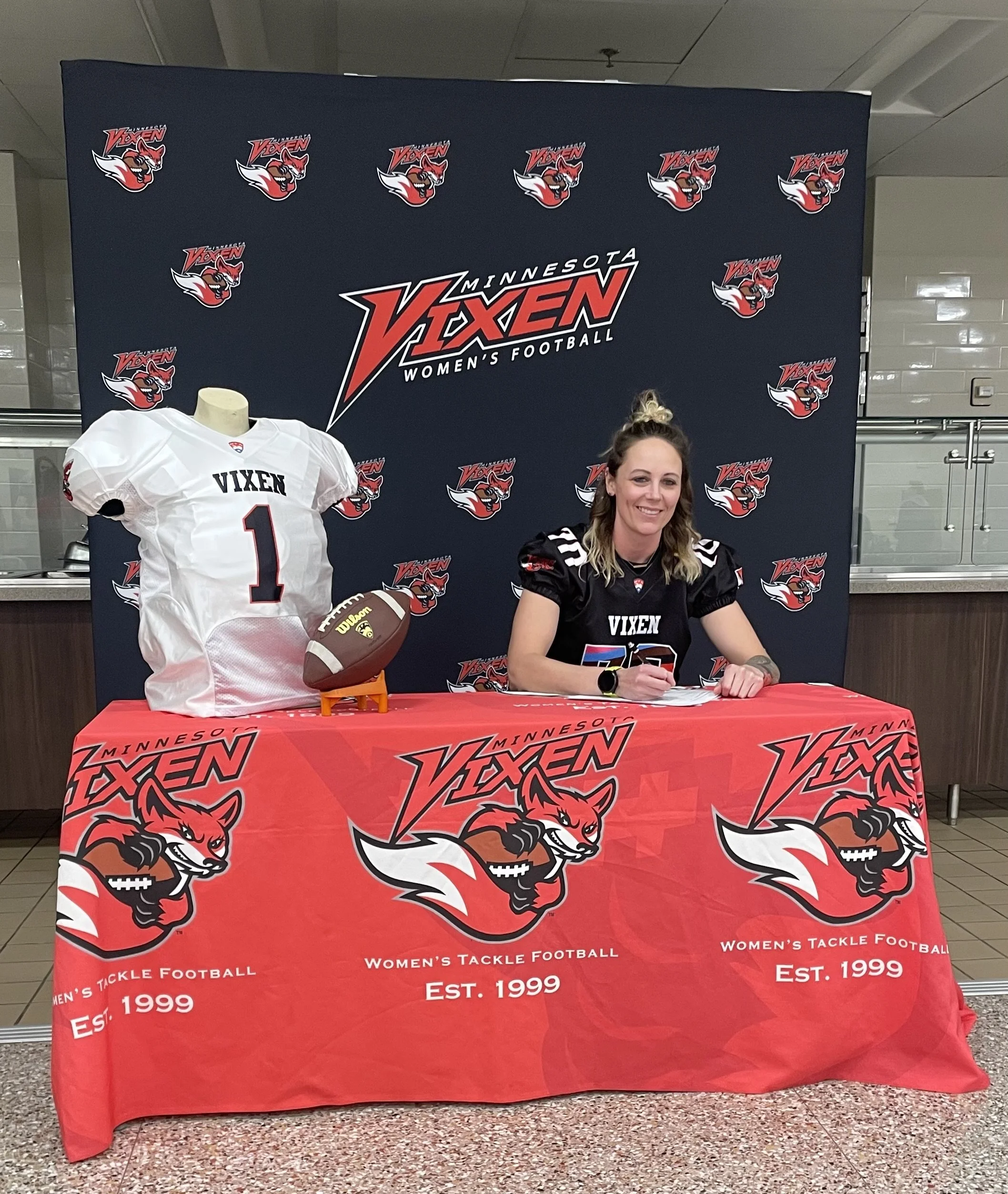 A woman in Minnesotta Vikings women's football team jersey sitting at a signing table with team logo cover, with football and jersey displayed next to her, in front of a Vixen women's football backdrop.