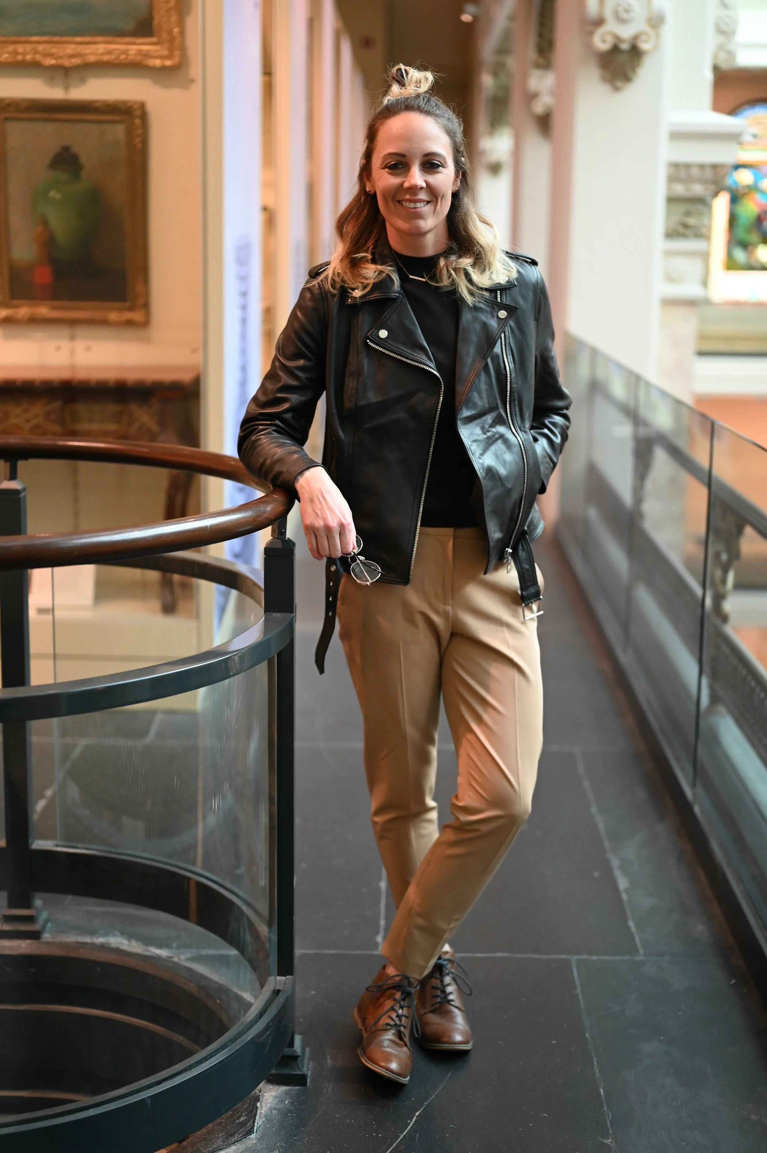 A woman with blonde hair in a bun, dressed in a black leather jacket, black top, beige pants, and brown boots, standing indoors in front of framed artwork, smiling at the camera.