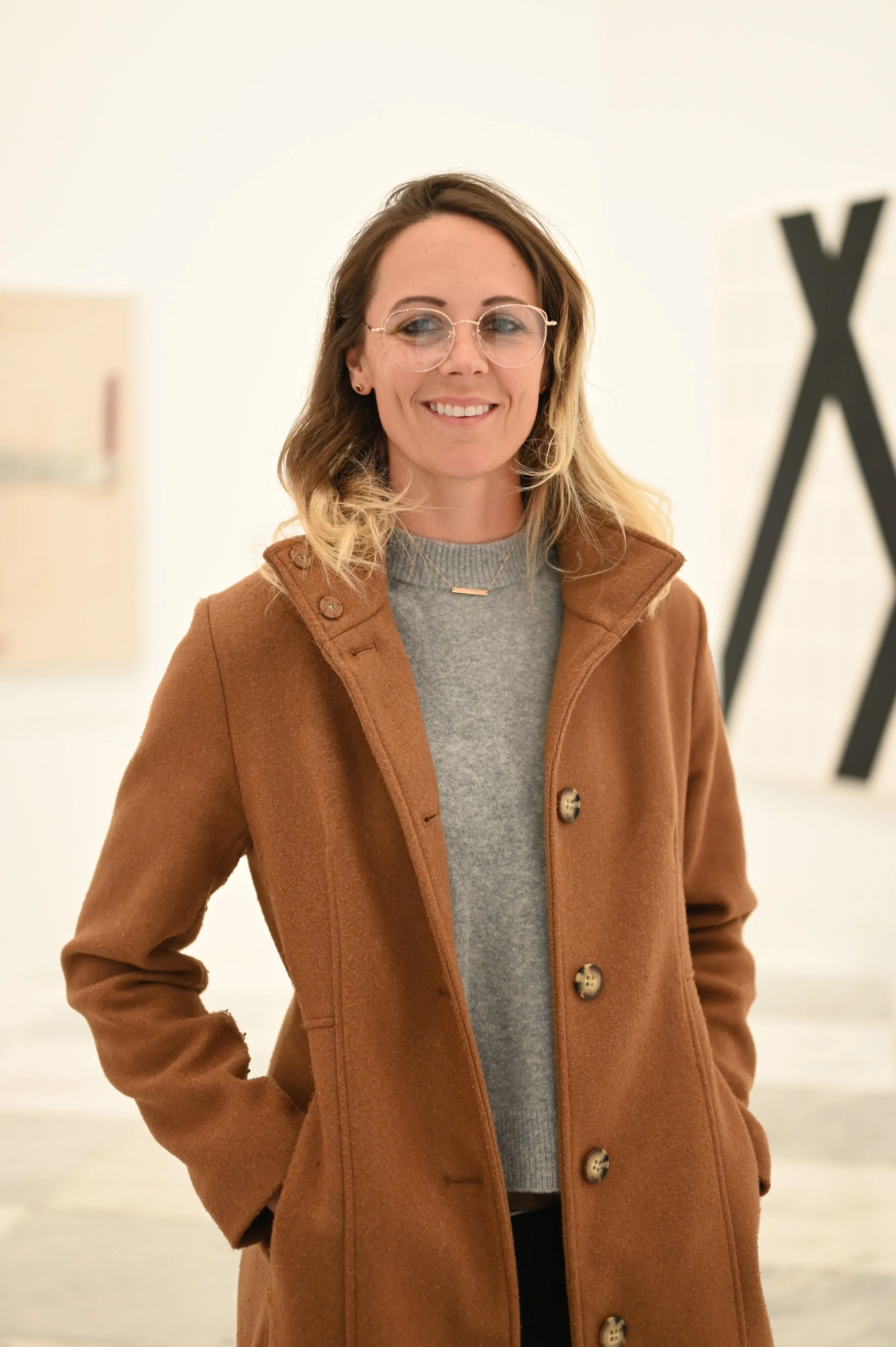 A woman with shoulder-length wavy hair, wearing glasses, a gray sweater, and a brown coat, standing indoors with hands in coat pockets, smiling at the camera.