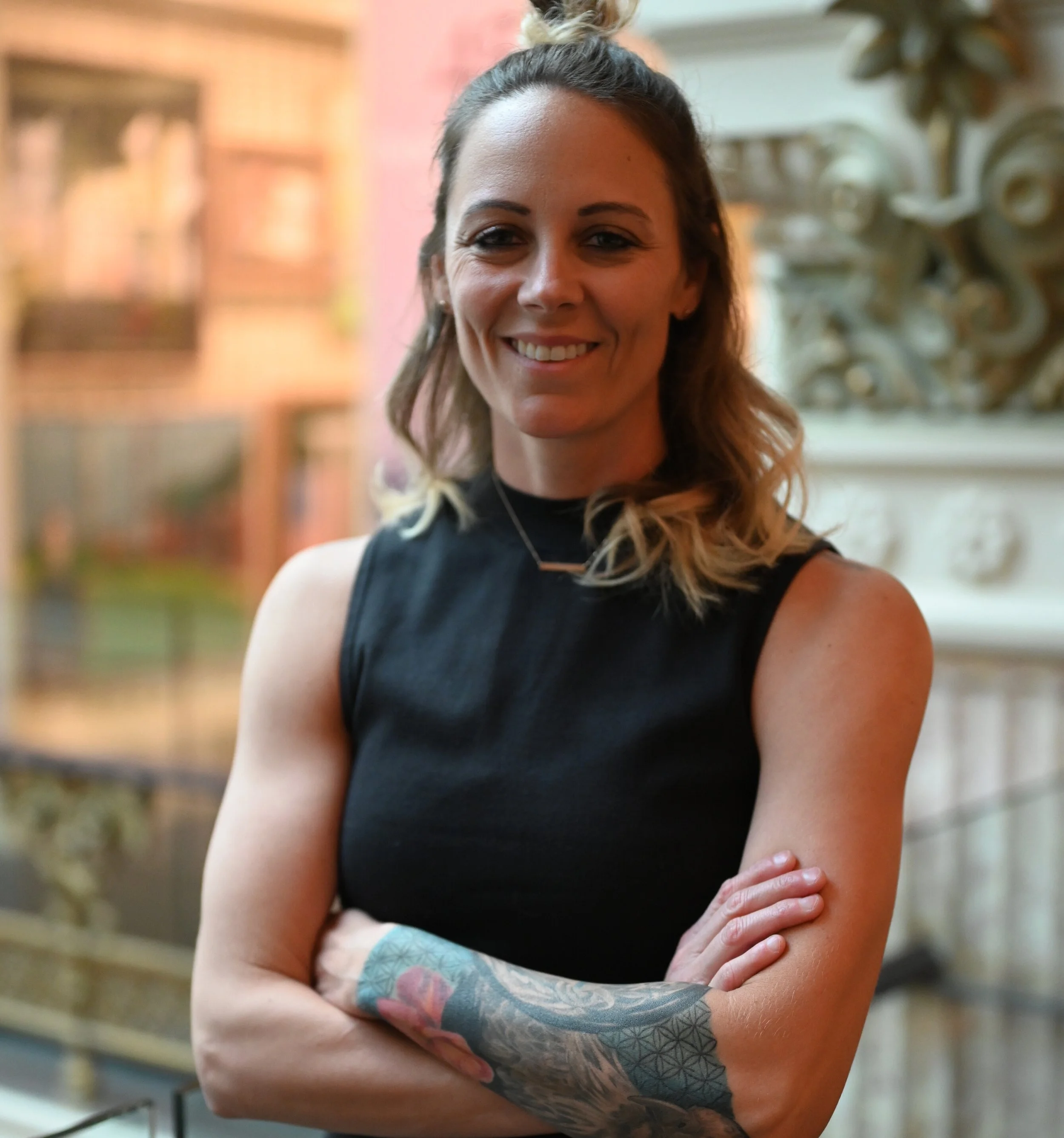 A smiling woman with wavy shoulder-length hair, wearing a sleeveless black top, standing indoors with her arms crossed, showing a colorful tattoo on her left forearm.