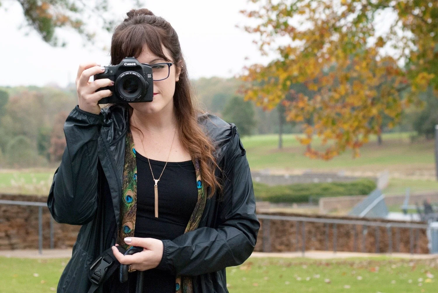 A woman with glasses and long brown hair taking a photo with a camera outdoors during autumn, wearing a black jacket and a black top, with colorful trees in the background.