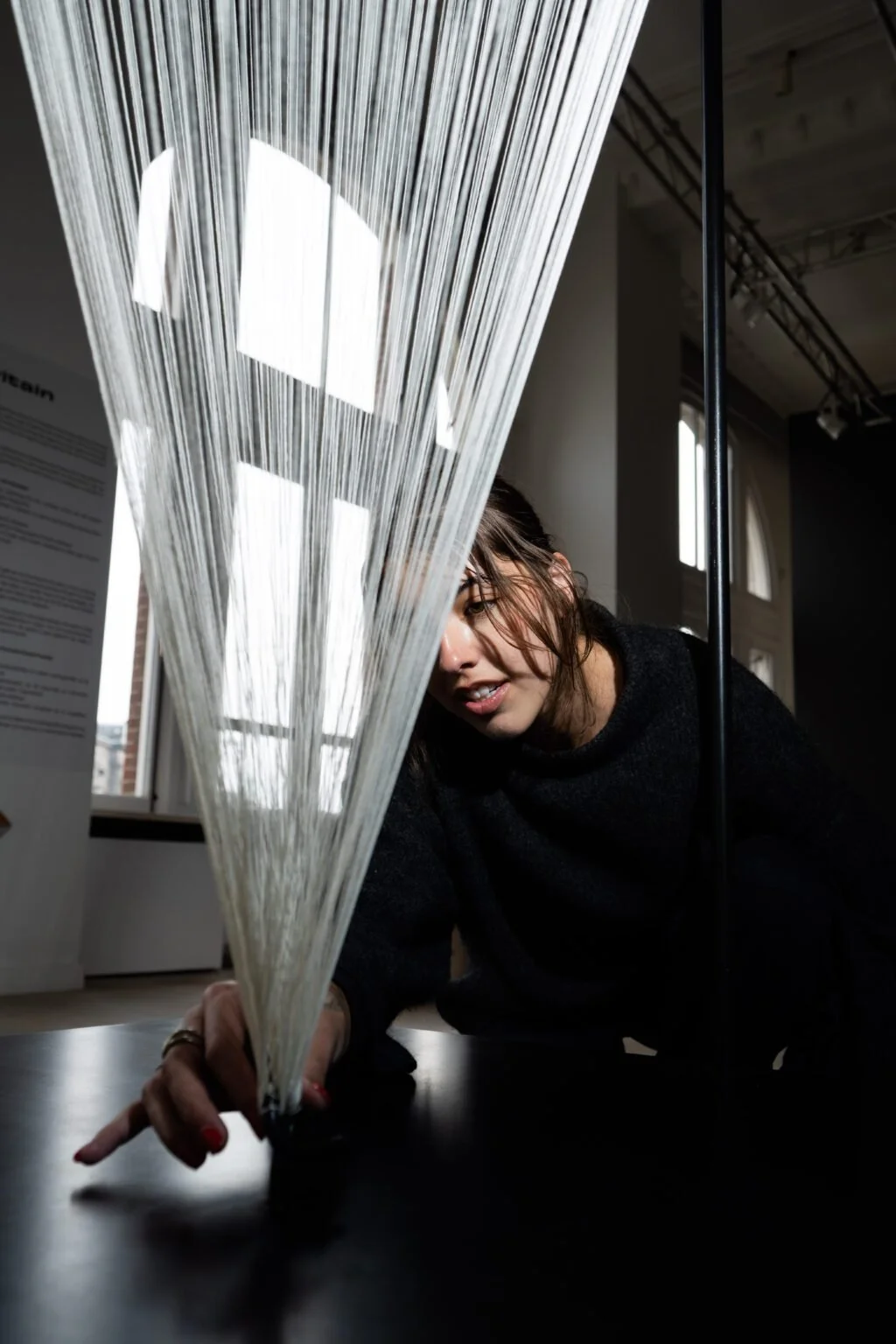 A woman leaning over a table, reaching through a hanging art installation made of strings, with windows and a display board in the background.
