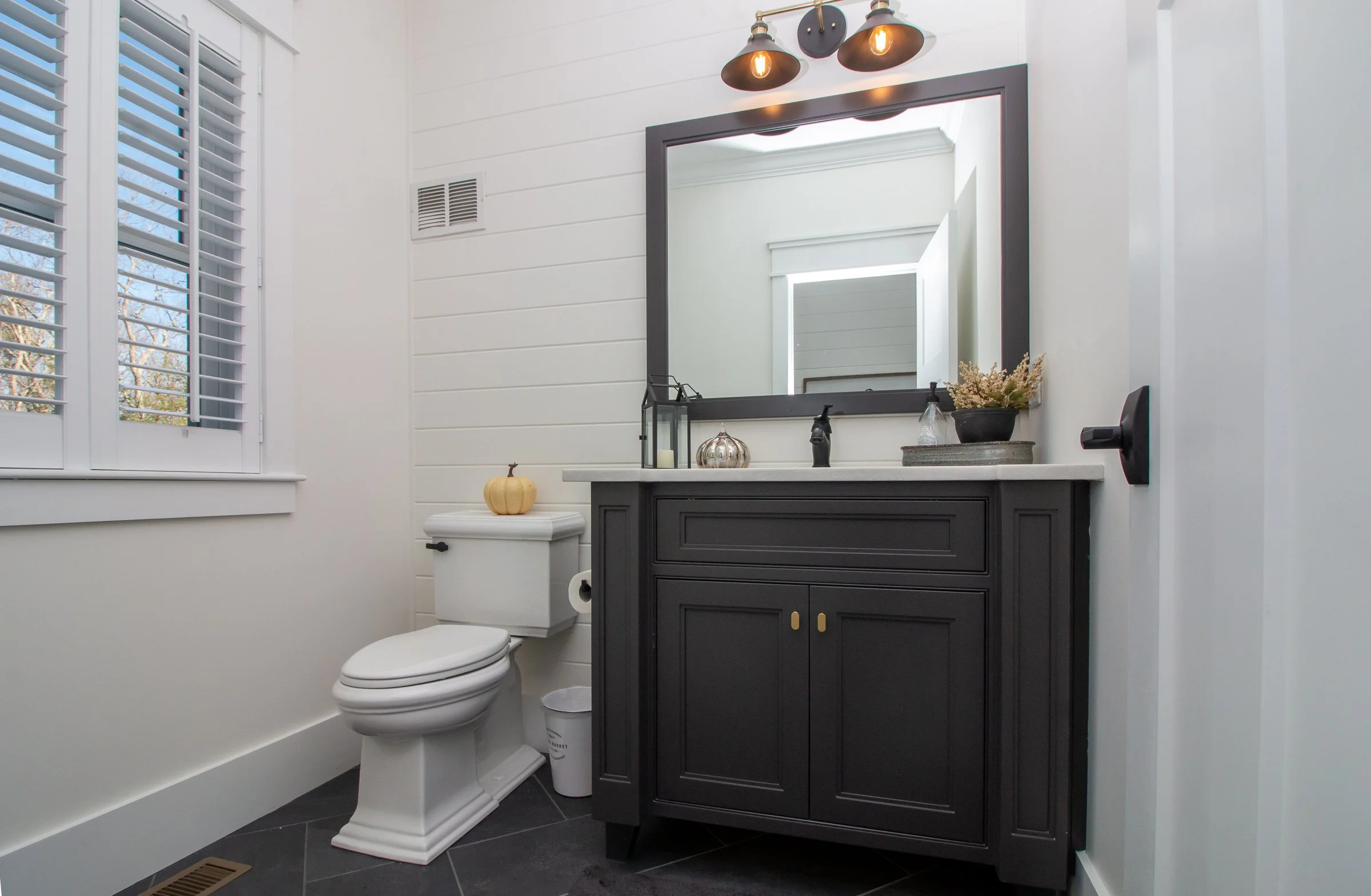 A modern bathroom with white walls, a window with shutters, a black vanity with a mirror, decorative lighting, and bathroom accessories, including a pumpkin on the toilet tank.