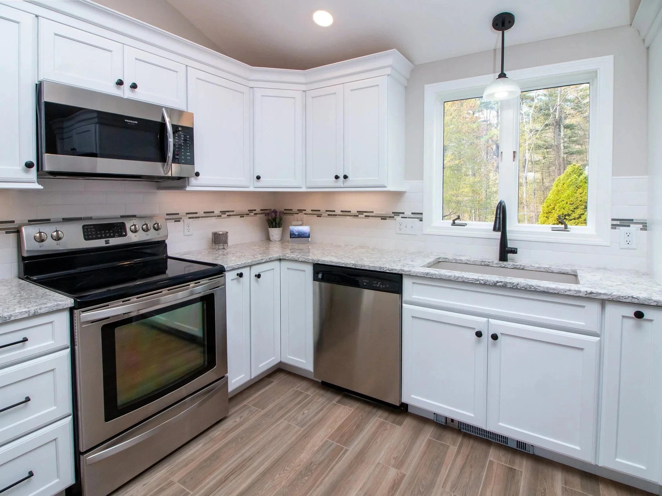 Modern kitchen with white cabinets, stainless steel stove, microwave, dishwasher, granite countertops, and a window overlooking trees.
