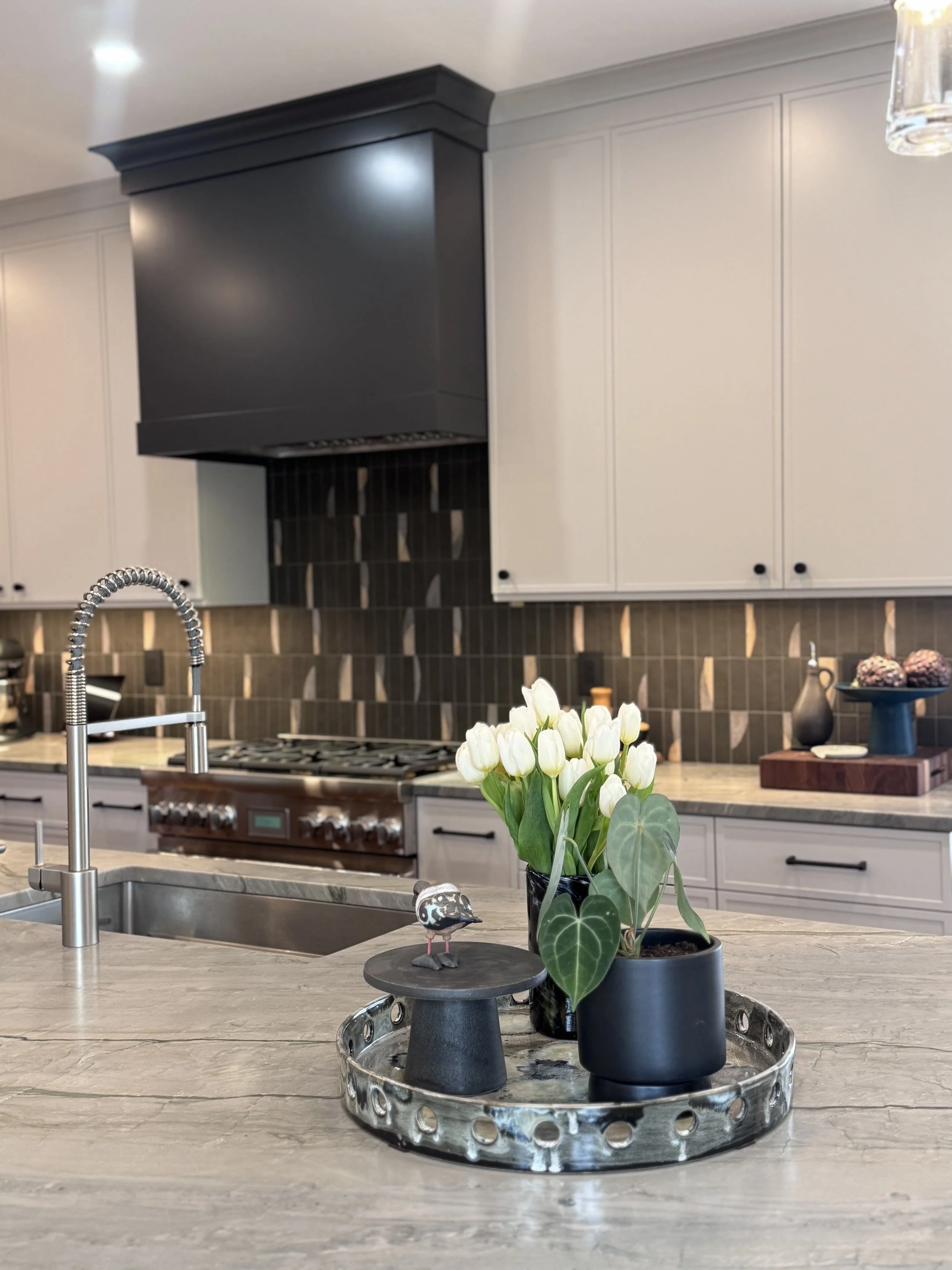 Kitchen with black range hood, white cabinets, gray marble countertop, vase with white tulips, and a decorative bird figurine on a tray.