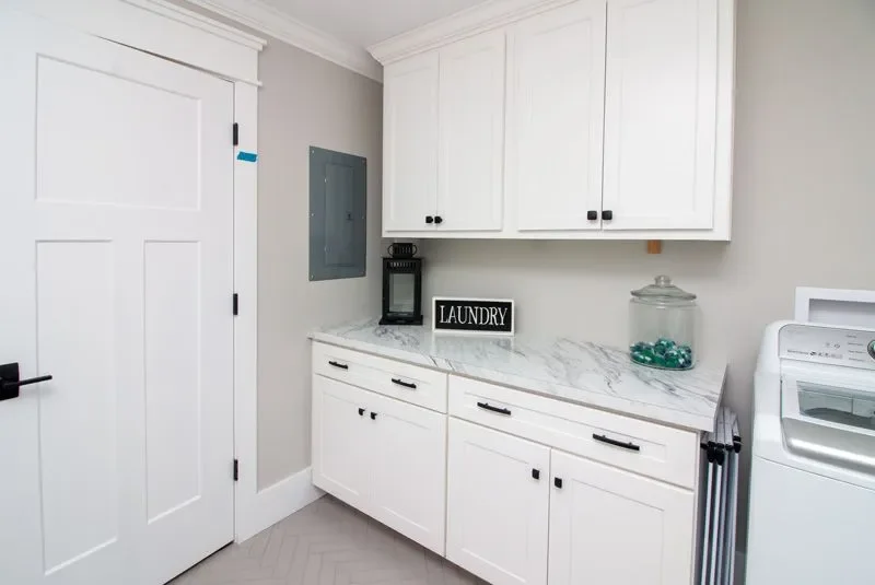 Laundry room with white cabinets, marble countertop, laundry sign, black coffee maker, a jar with cleaning tabs, and a washing machine.