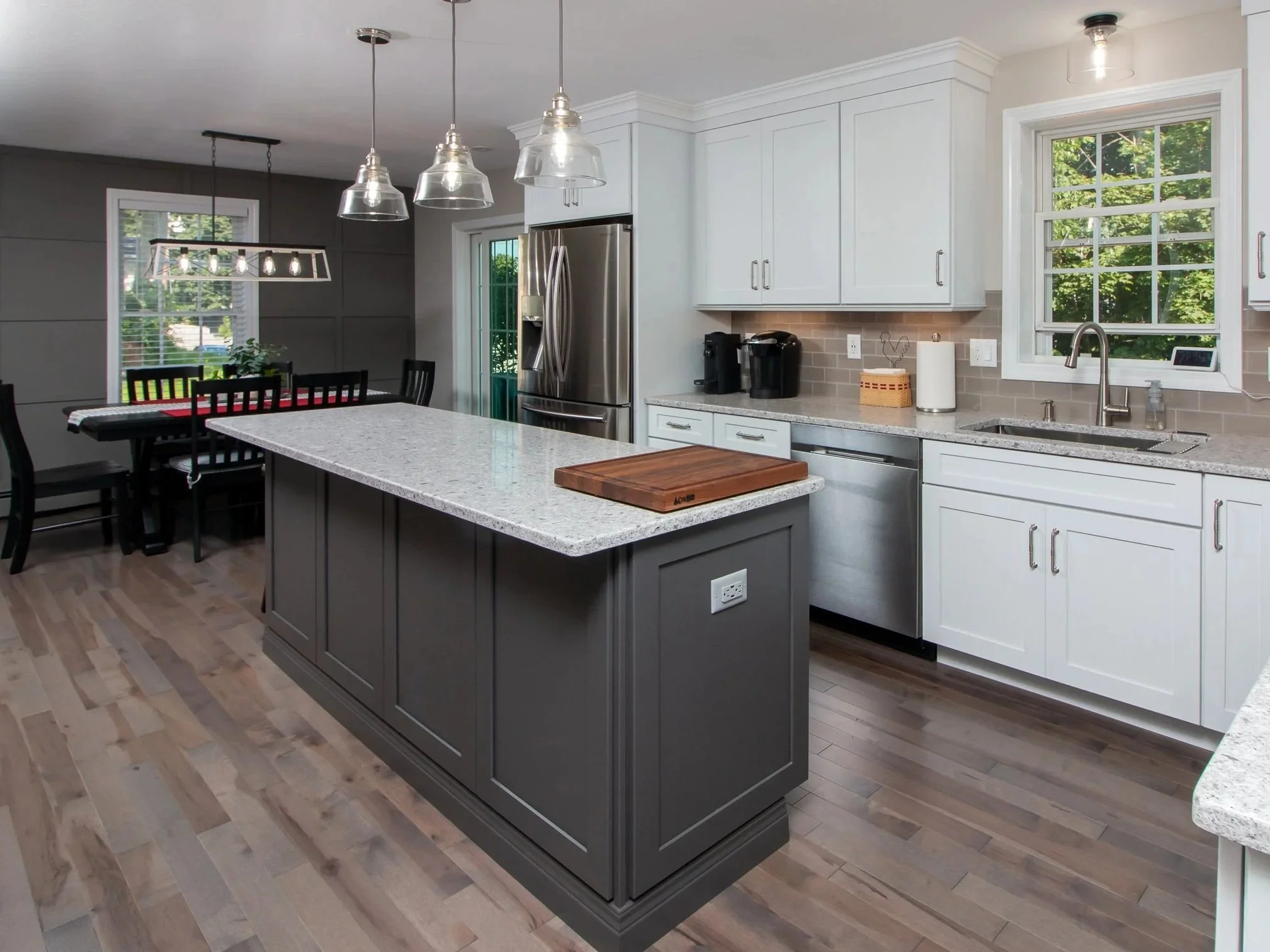Modern kitchen with white cabinets, stainless steel appliances, a dark gray island with a granite countertop, and a dining area with black chairs and a table. Light hardwood floors and windows with greenery outside.