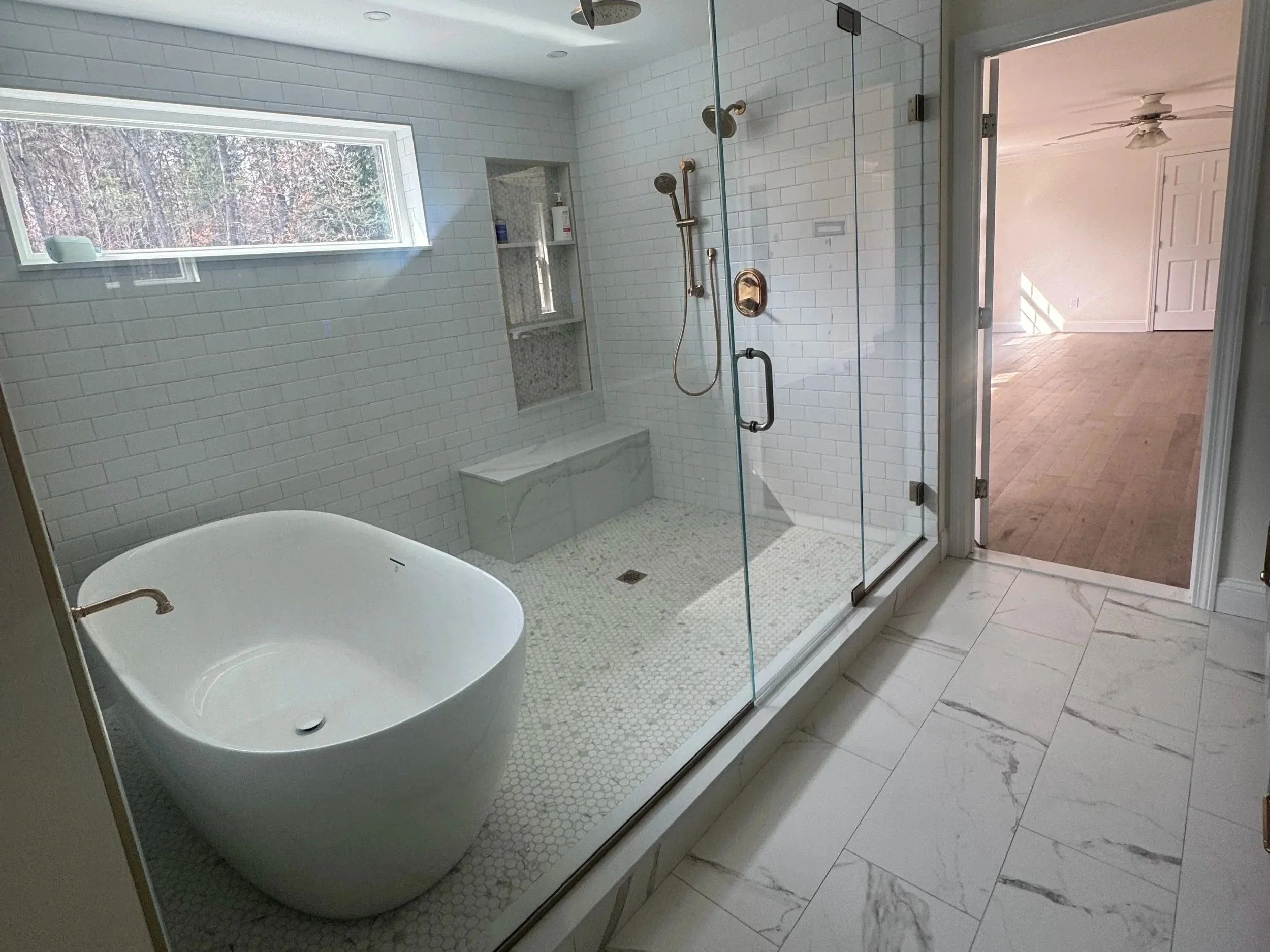 Modern bathroom with a glass-enclosed shower and a freestanding bathtub, white brick wall tiles, and a large window showing trees outside.