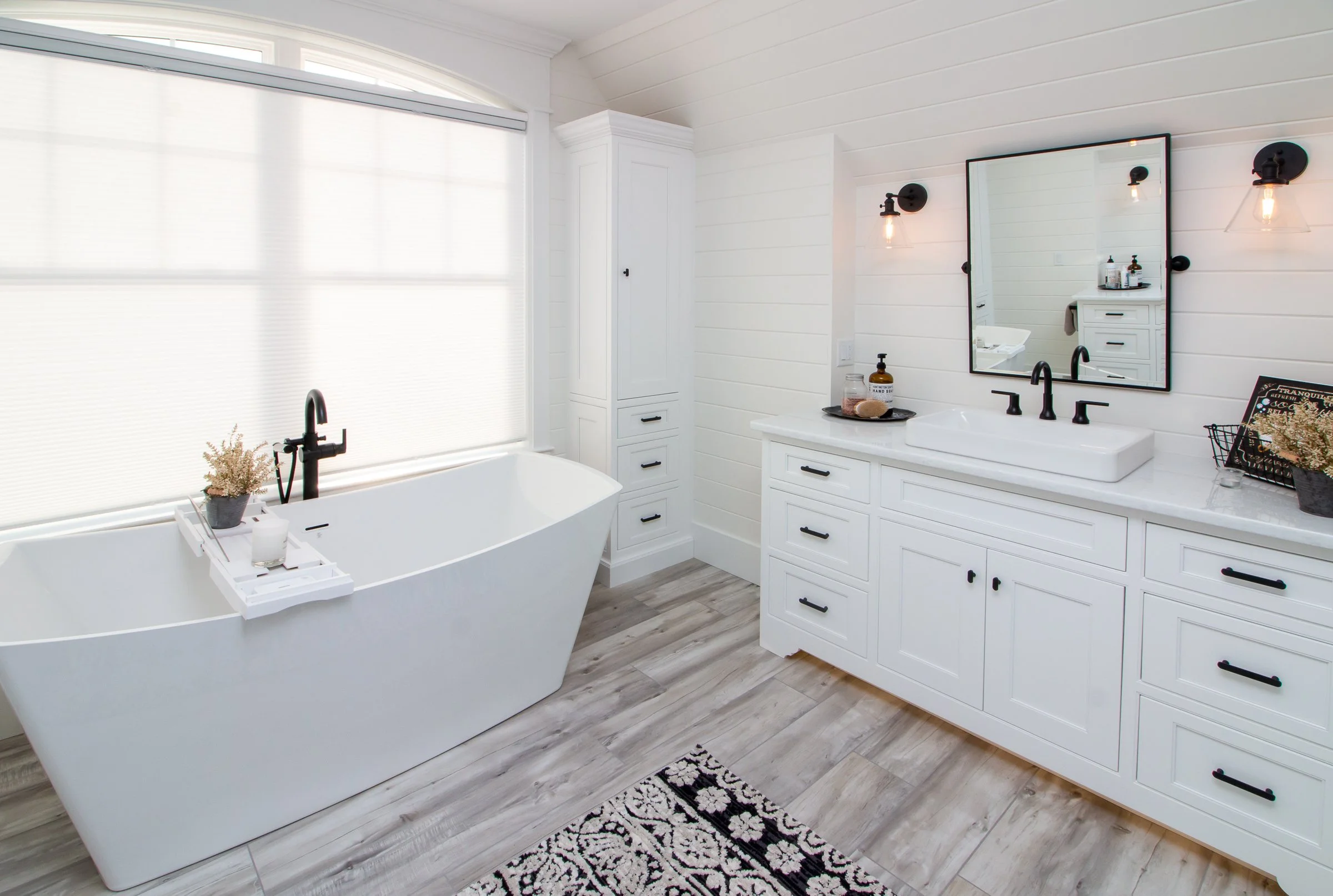 Modern white bathroom with a large bathtub next to frosted windows, white cabinetry, and a double sink vanity with black fixtures and a mirror. Decor includes a black and white rug and small plants.