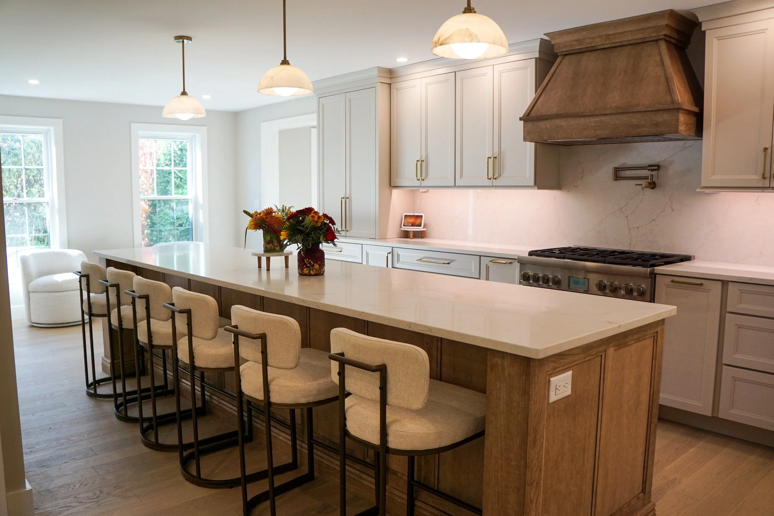 Modern kitchen with white cabinetry, marble backsplash, large island with beige counter, floral centerpiece, pendant lights, and stainless steel stove.