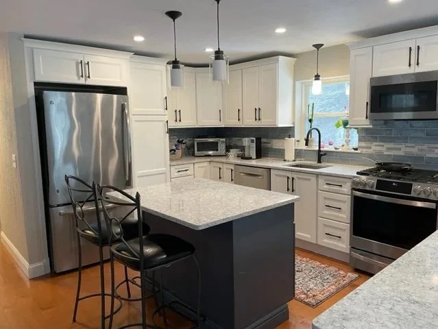 Modern kitchen with white cabinets, stainless steel appliances, a kitchen island with two black bar stools, pendant lights, and a window above the sink.