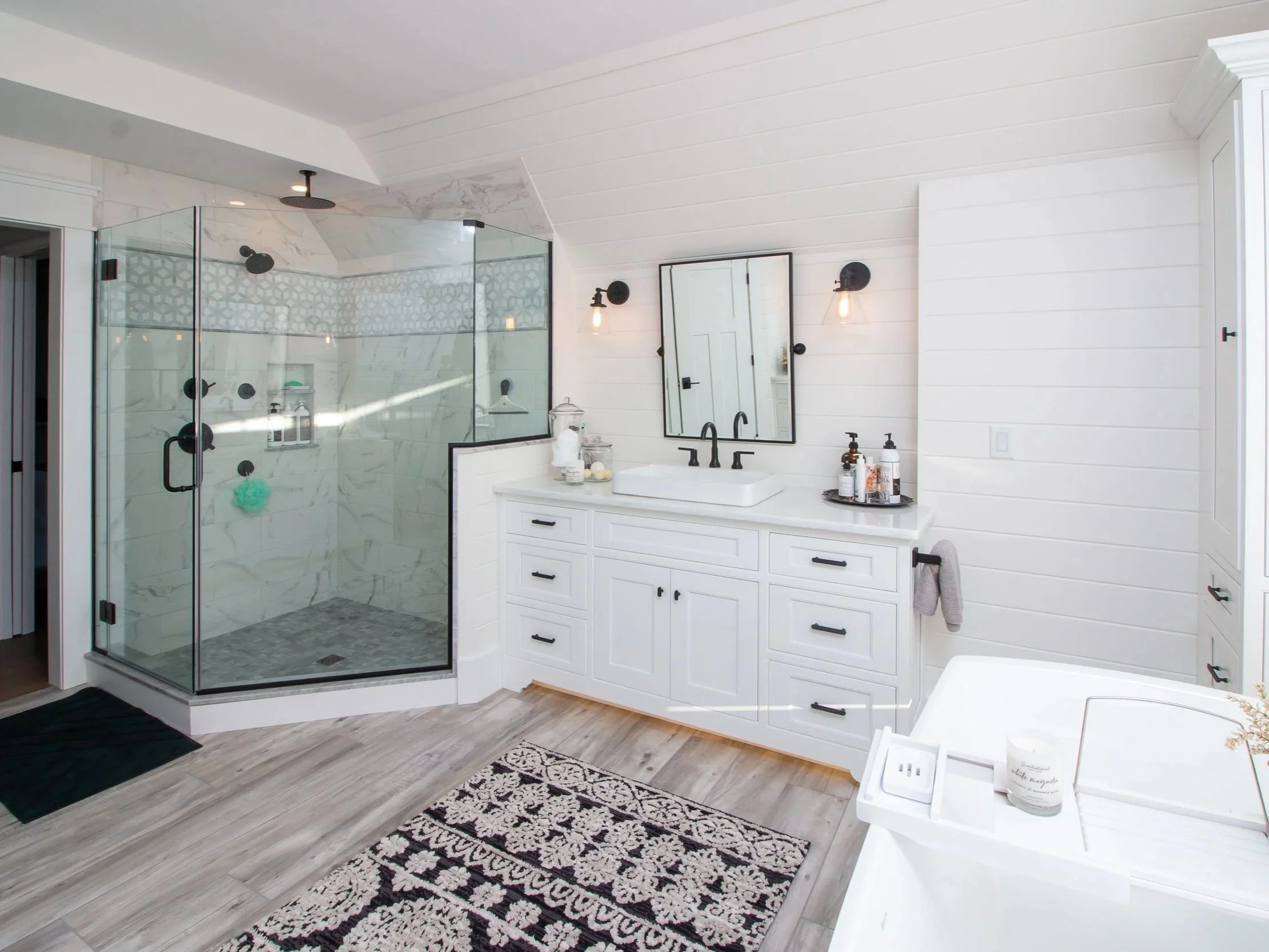 Modern white bathroom with a glass shower enclosure, white vanity with black hardware, wall-mounted mirror, wall-mounted light fixtures, a bathtub, and wooden flooring.