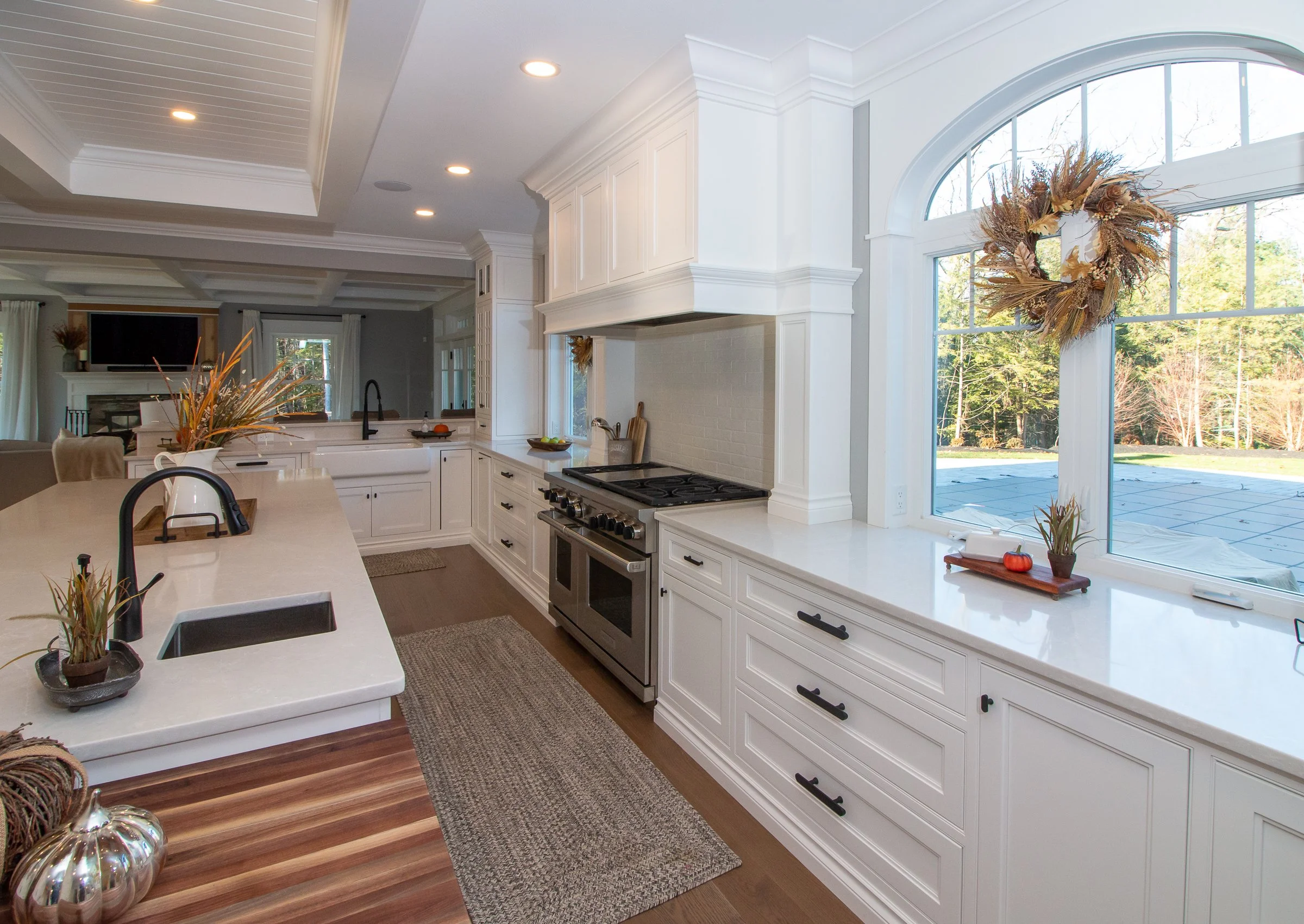 Bright kitchen with white cabinets, a large window with a fall wreath, and autumn decorations, including pumpkins and dried plants, on the window sill and countertop.