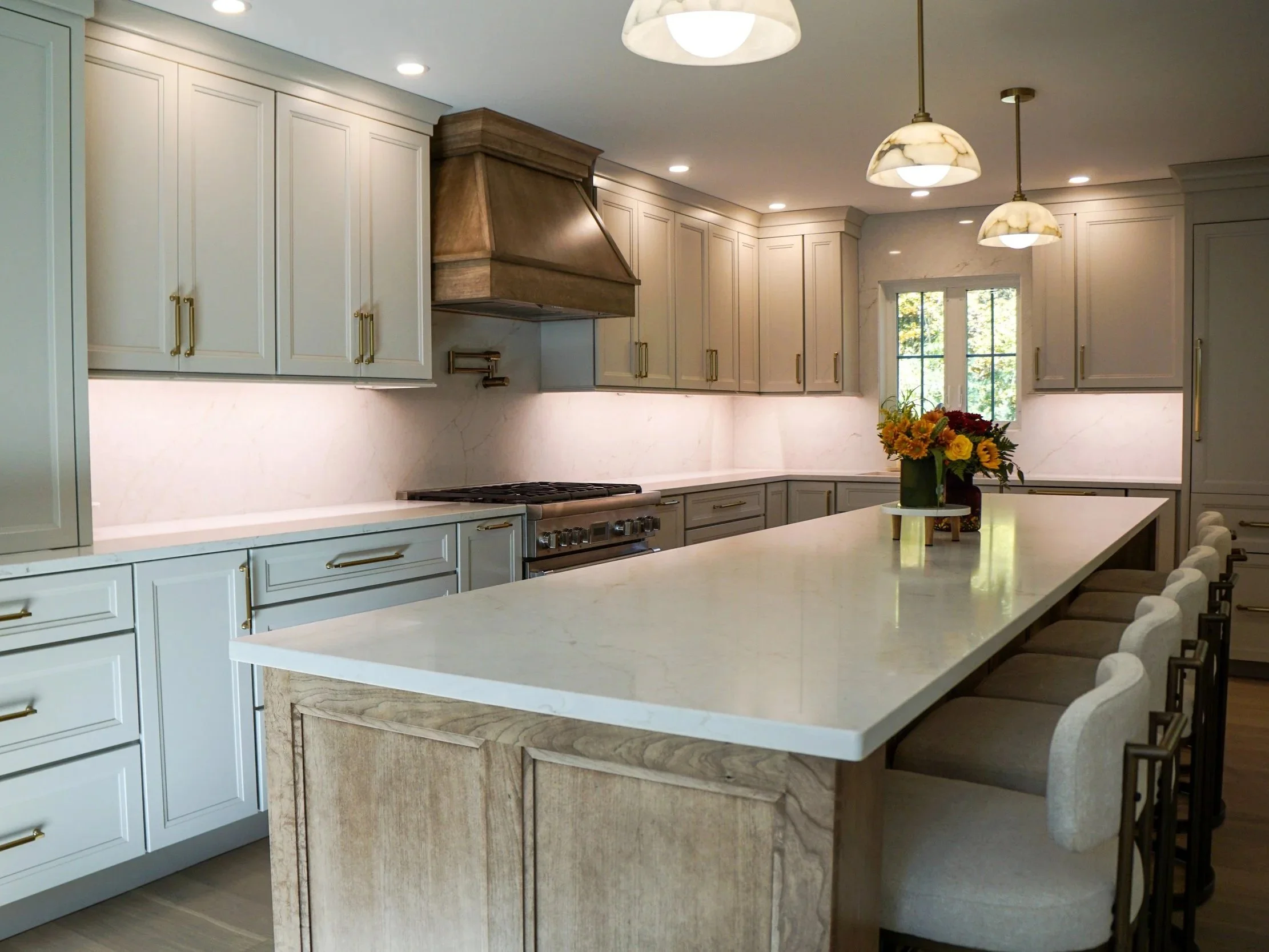 Modern kitchen with white and gray cabinets, a large island with a white countertop, a vase of colorful flowers, decorated with pendant lights and a window showing greenery outside.