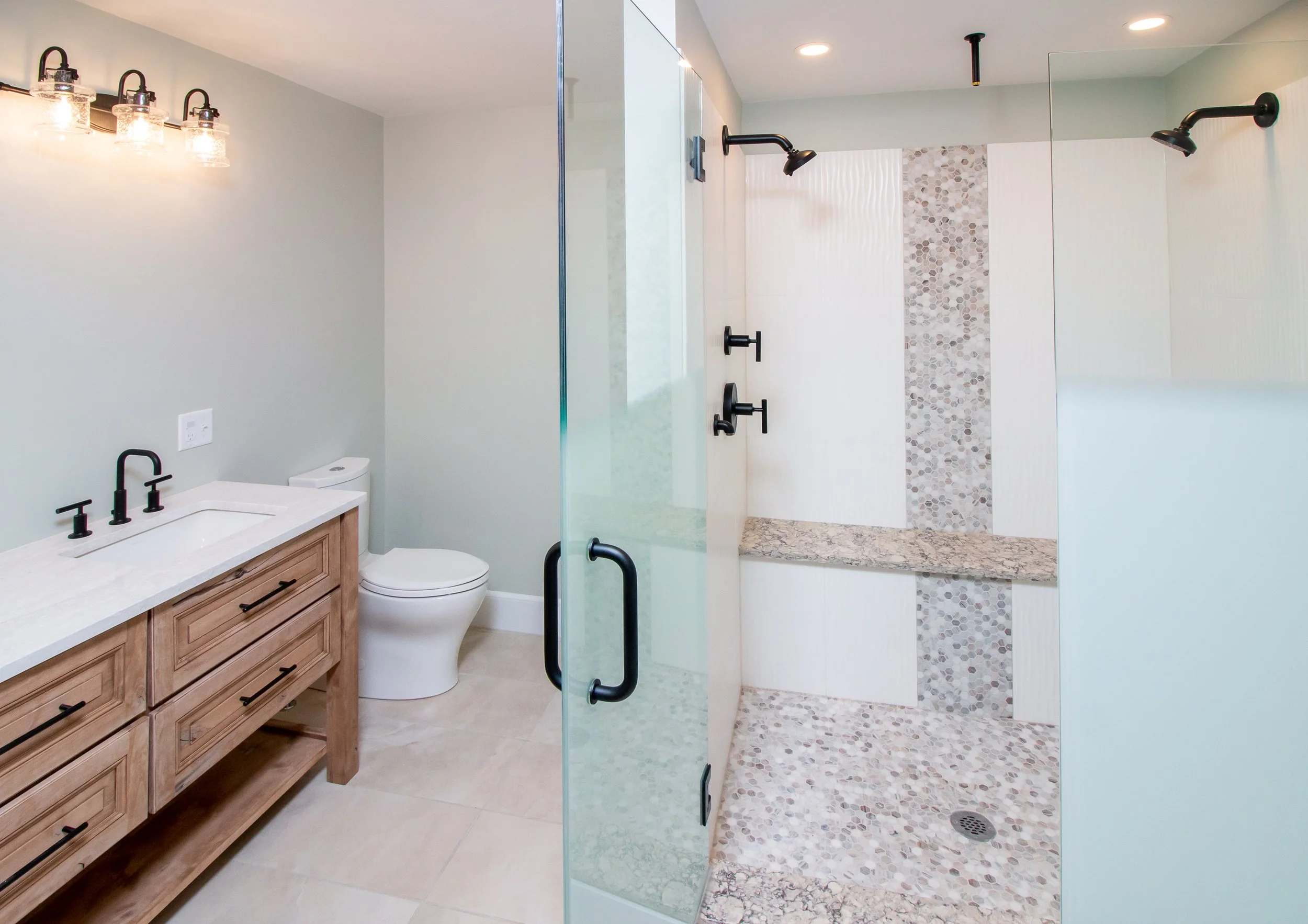 Modern bathroom with a wooden vanity, white countertop, black faucet, toilet, and a walk-in shower with glass enclosure, black fixtures, and stone tile accents.