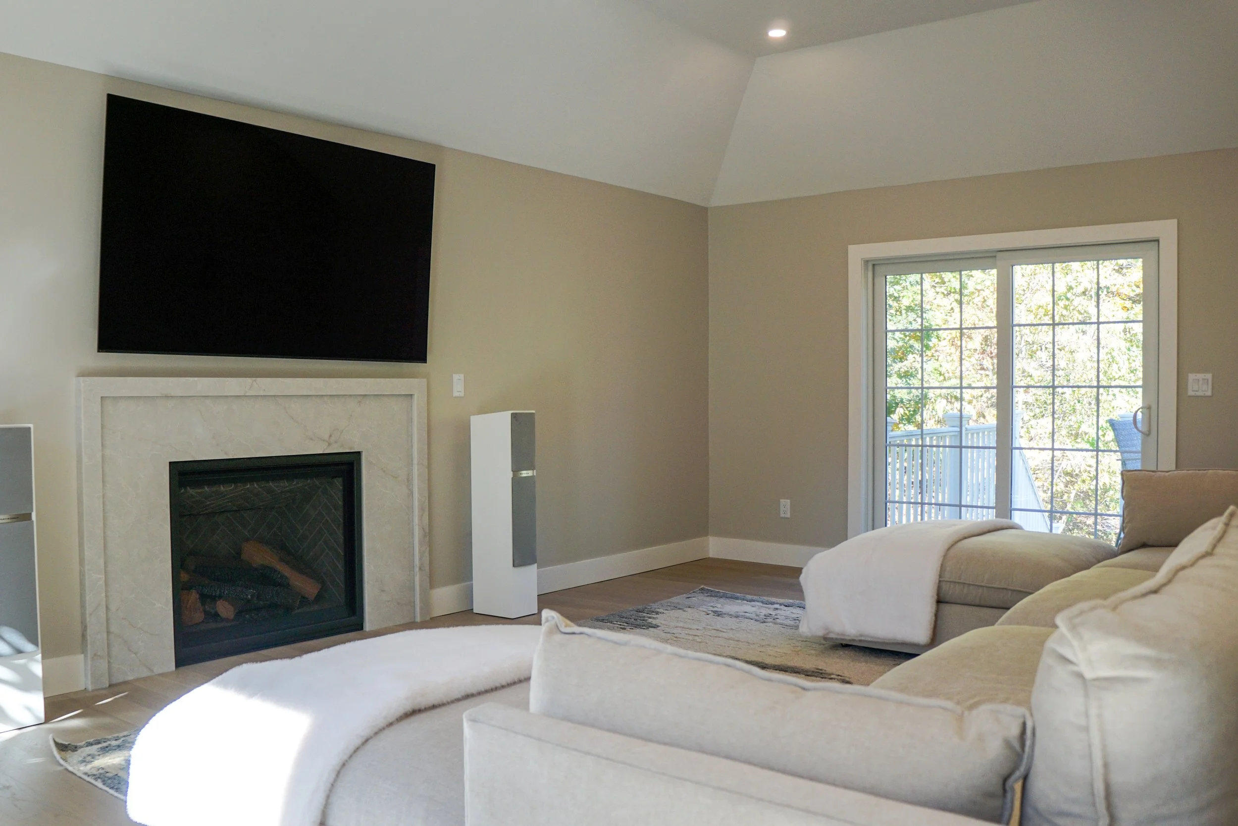 Living room with beige sofas, a fireplace, a large TV above the fireplace, and a sliding glass door leading outside to a deck with trees visible.
