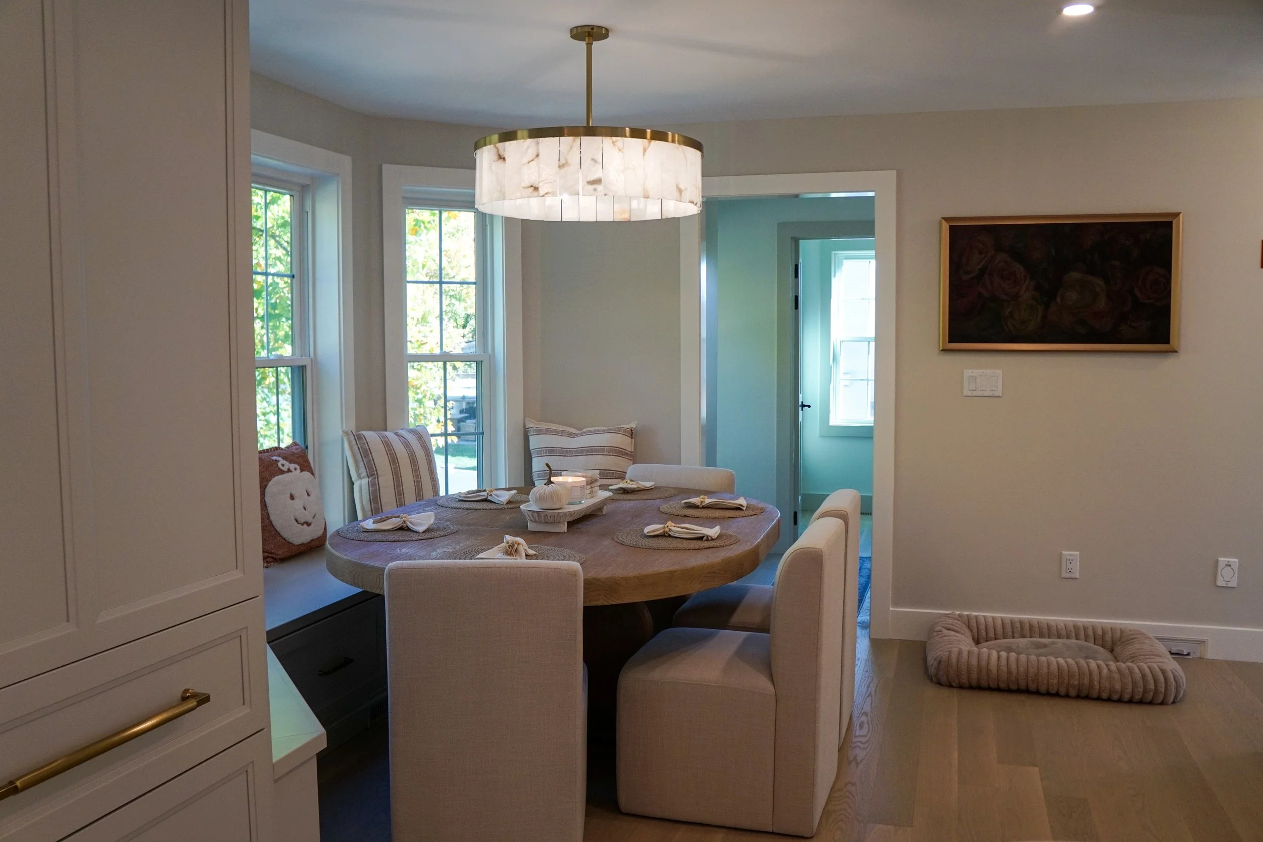 A cozy dining area with a round wooden table set with placemats and napkins, surrounded by upholstered chairs. There is a window seat with throw pillows, a window with a view of greenery, a modern pendant light overhead, and a wall with framed artwork. A pet bed is on the floor near the wall.