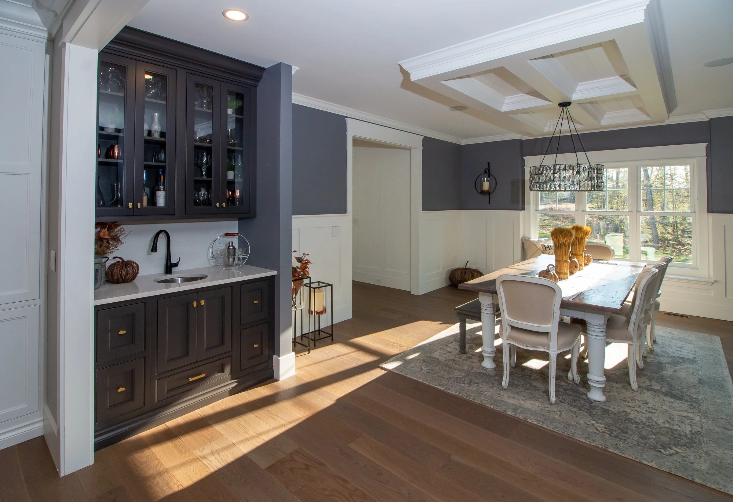 Dining room with large window, white and dark gray walls, wooden dining table with six white chairs, decorative vases, chandelier, and built-in cabinetry.
