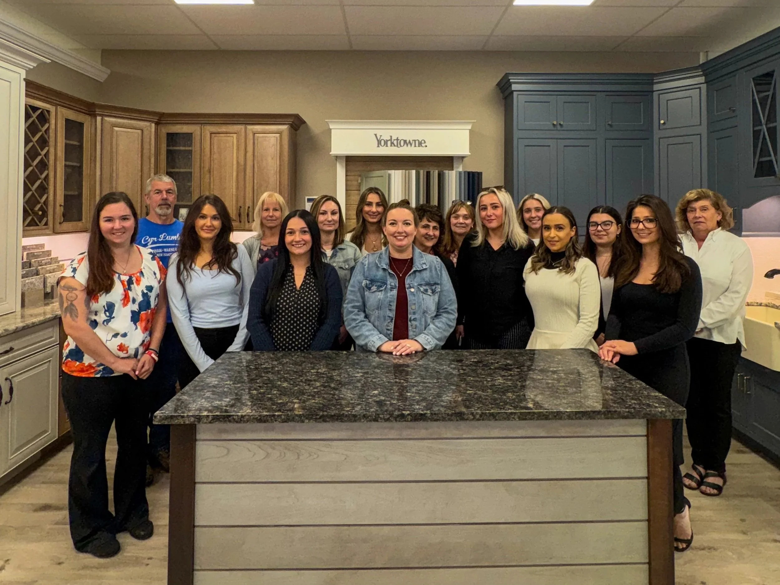 Group of 15 people standing in a kitchen showroom, posing for a photo behind a granite-topped island.