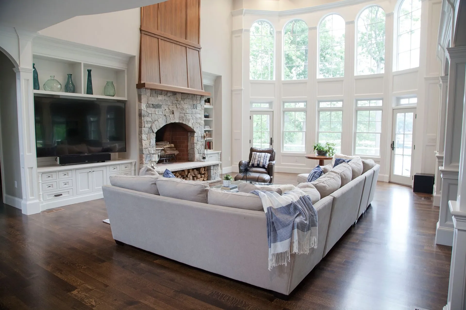 Living room with large bay windows, beige sectional sofa with blue and white pillows, leather armchair, stone fireplace, flat-screen TV, wood flooring, and white built-in shelves.