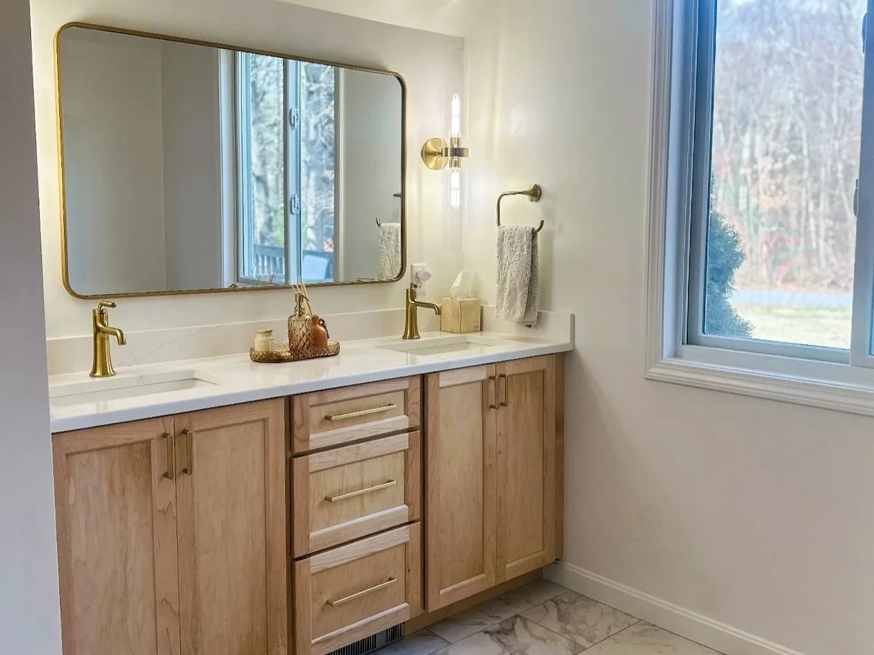 Modern bathroom vanity with light wood cabinets, a white marble countertop, and gold fixtures, includes a large mirror, a window, and decorative items.