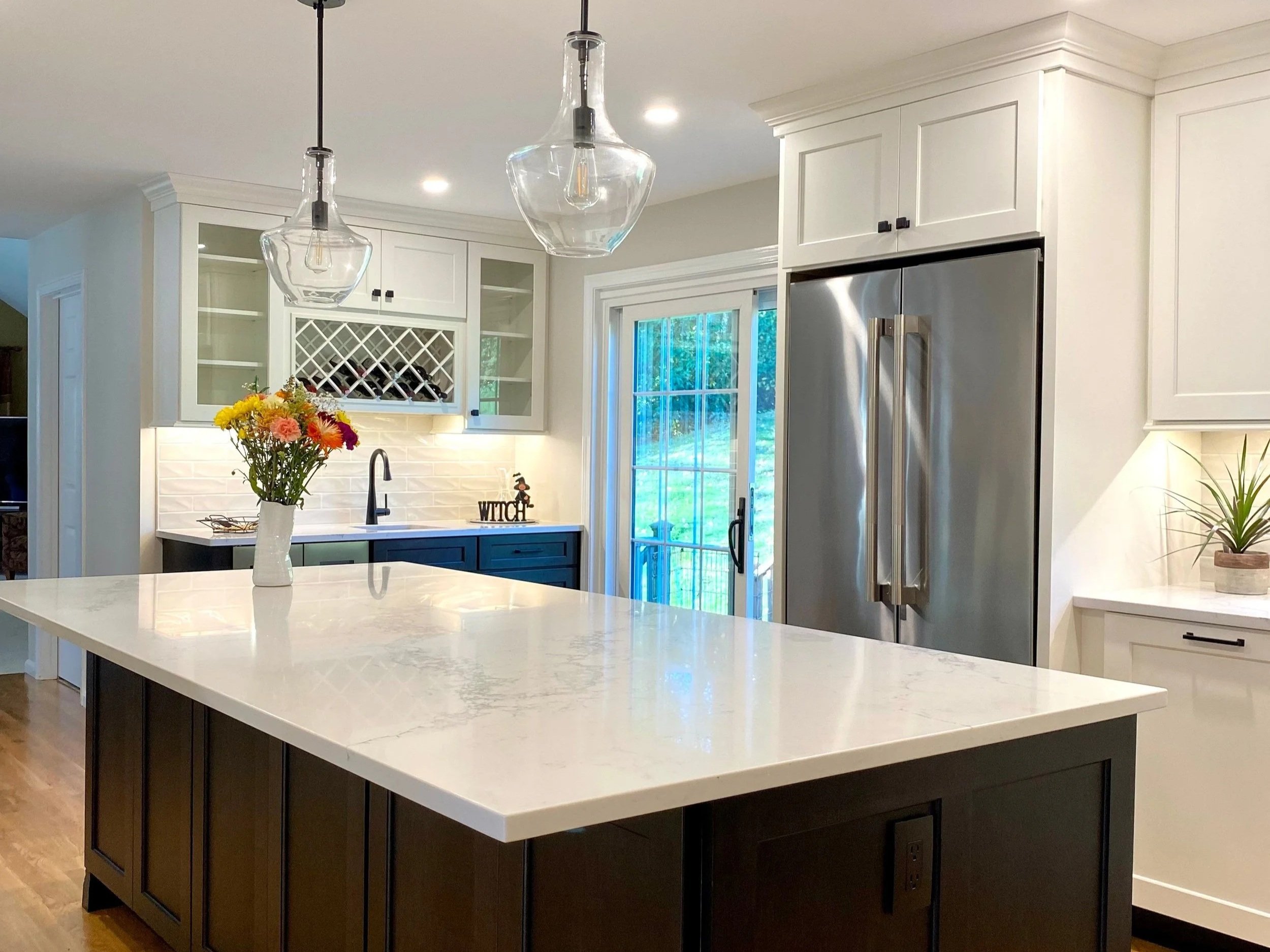 Modern kitchen with white cabinets, stainless steel refrigerator, black countertops, and an island with a white marble top. Decor includes a vase of flowers and pendant lights.