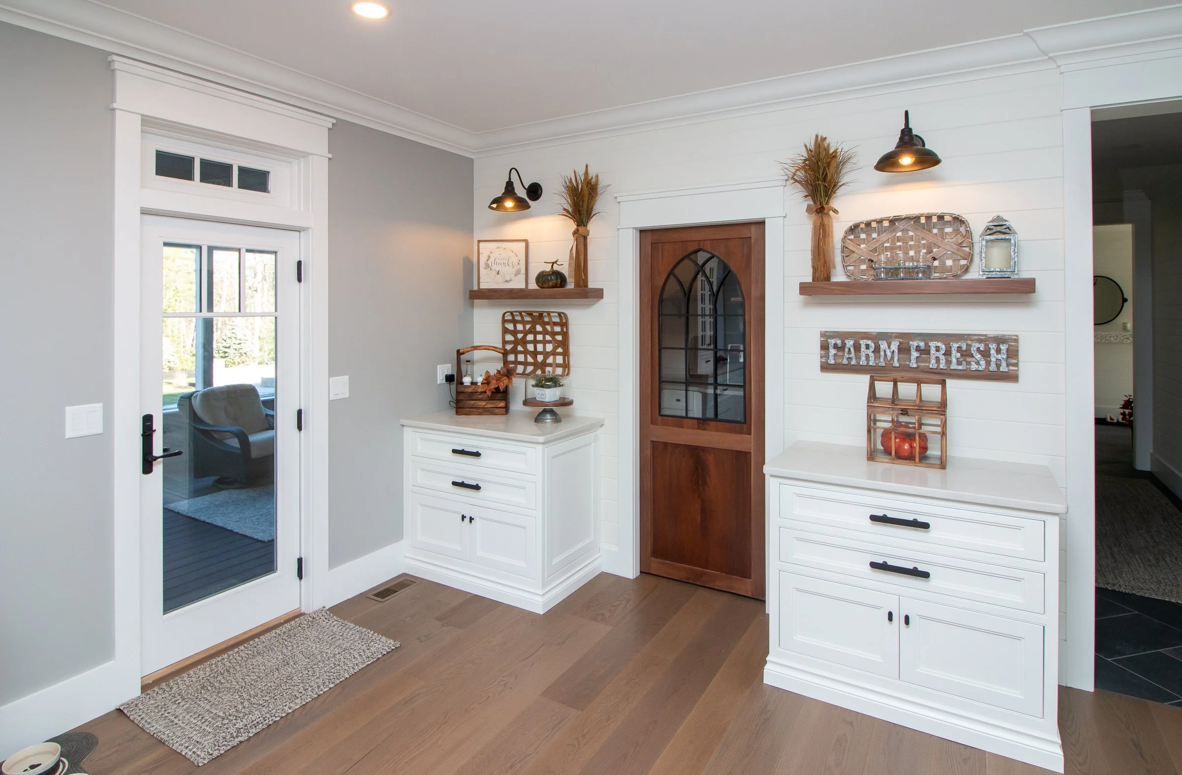 Interior entryway with white cabinetry, wooden accents, and farm-fresh decor, including shelves with vases, greenery, and a sign, and a brown door with glass panels.