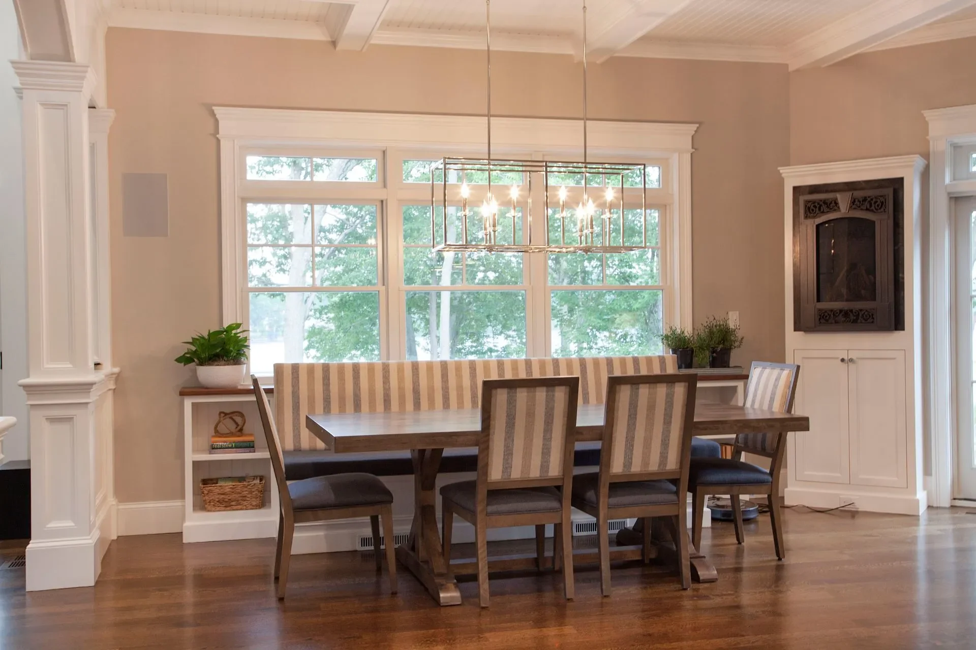 A dining room with a wooden table and six chairs, a built-in bench, large windows showing trees outside, and a modern chandelier hanging above the table.