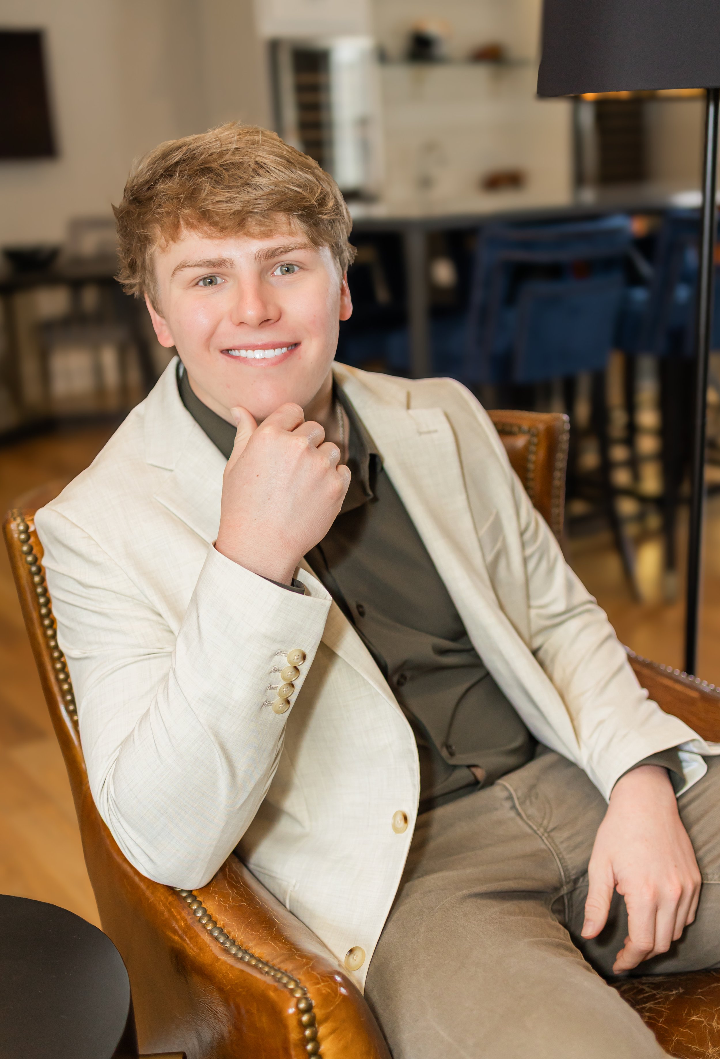A young man with short, curly blond hair and blue eyes, smiling, sitting in a brown leather chair in a modern living room or café. He is wearing a beige blazer over a dark shirt and has his right hand resting near his chin.