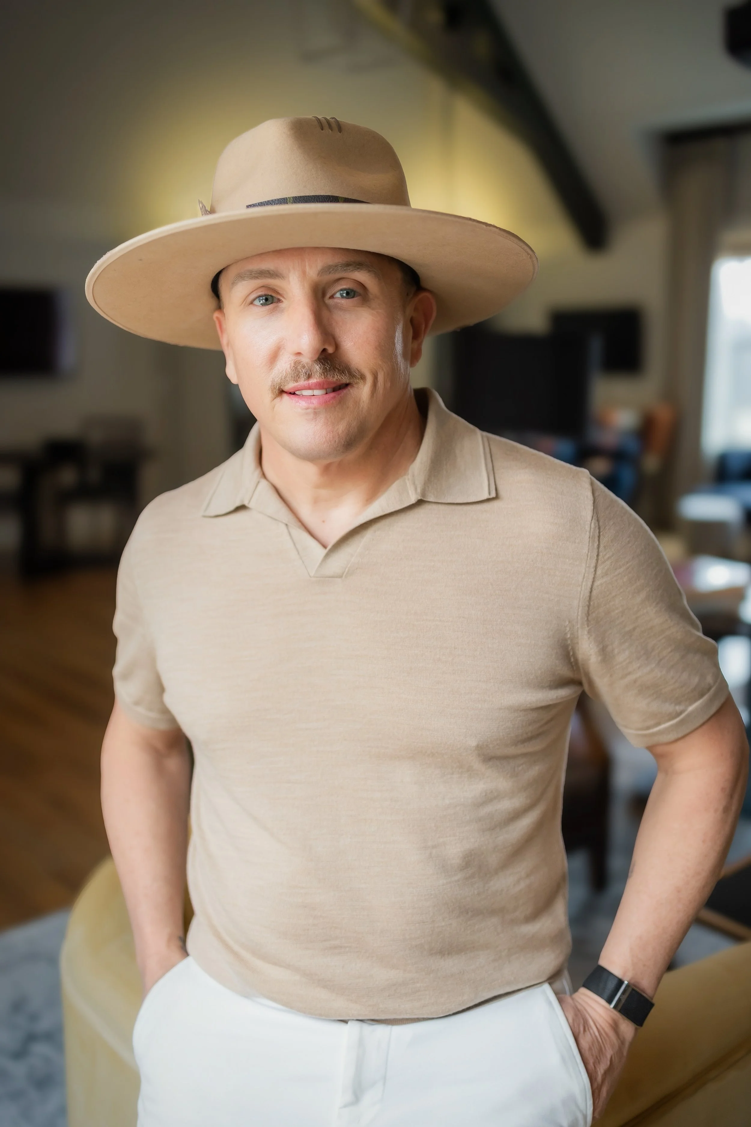 Man wearing a beige polo shirt, white pants, a large beige hat, and a black wristwatch, standing in a cozy, well-lit living room.